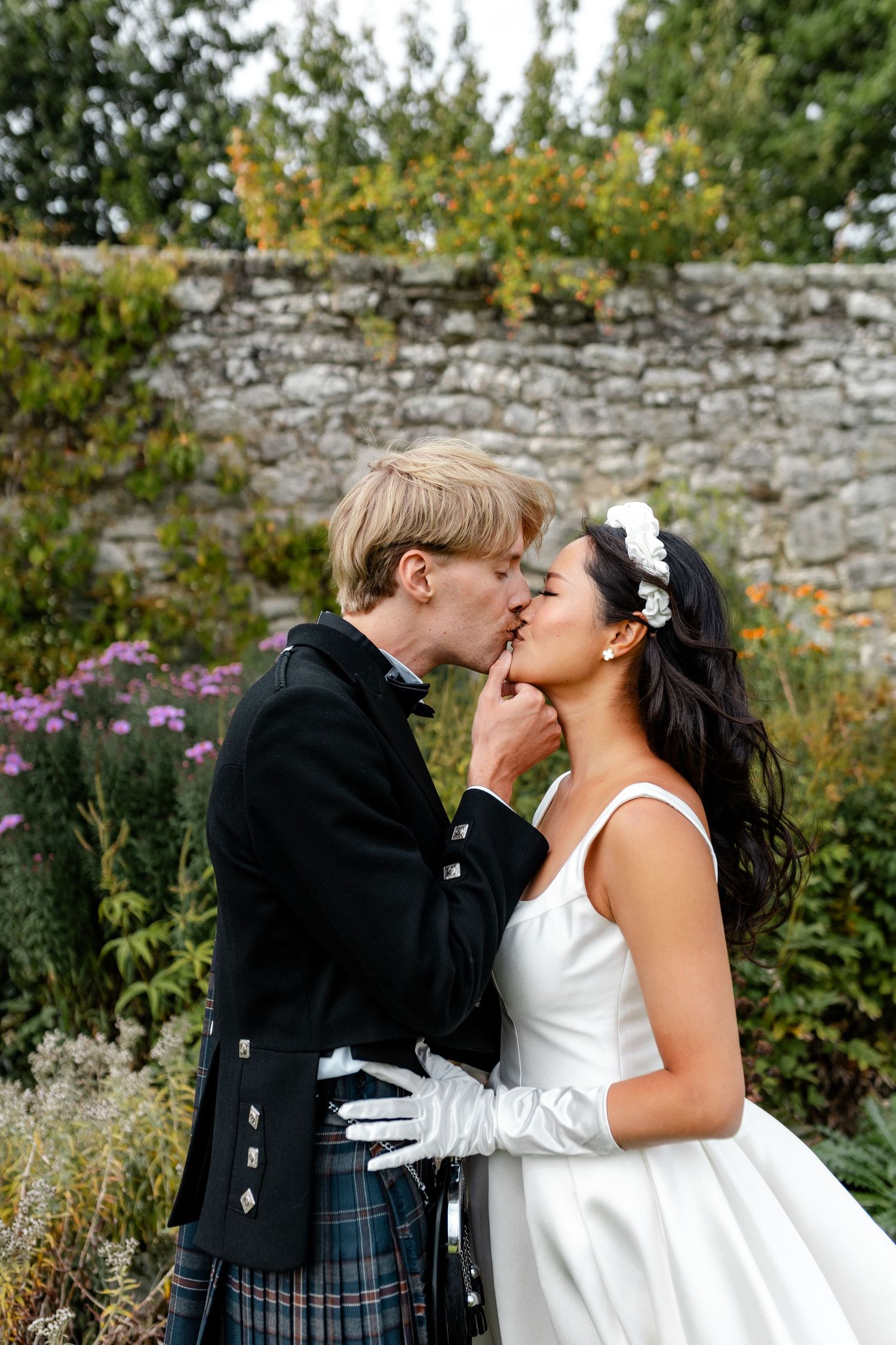 Bride and groom share a romantic kiss in a Scottish garden, groom wearing traditional tartan kilt and bride in white gown.