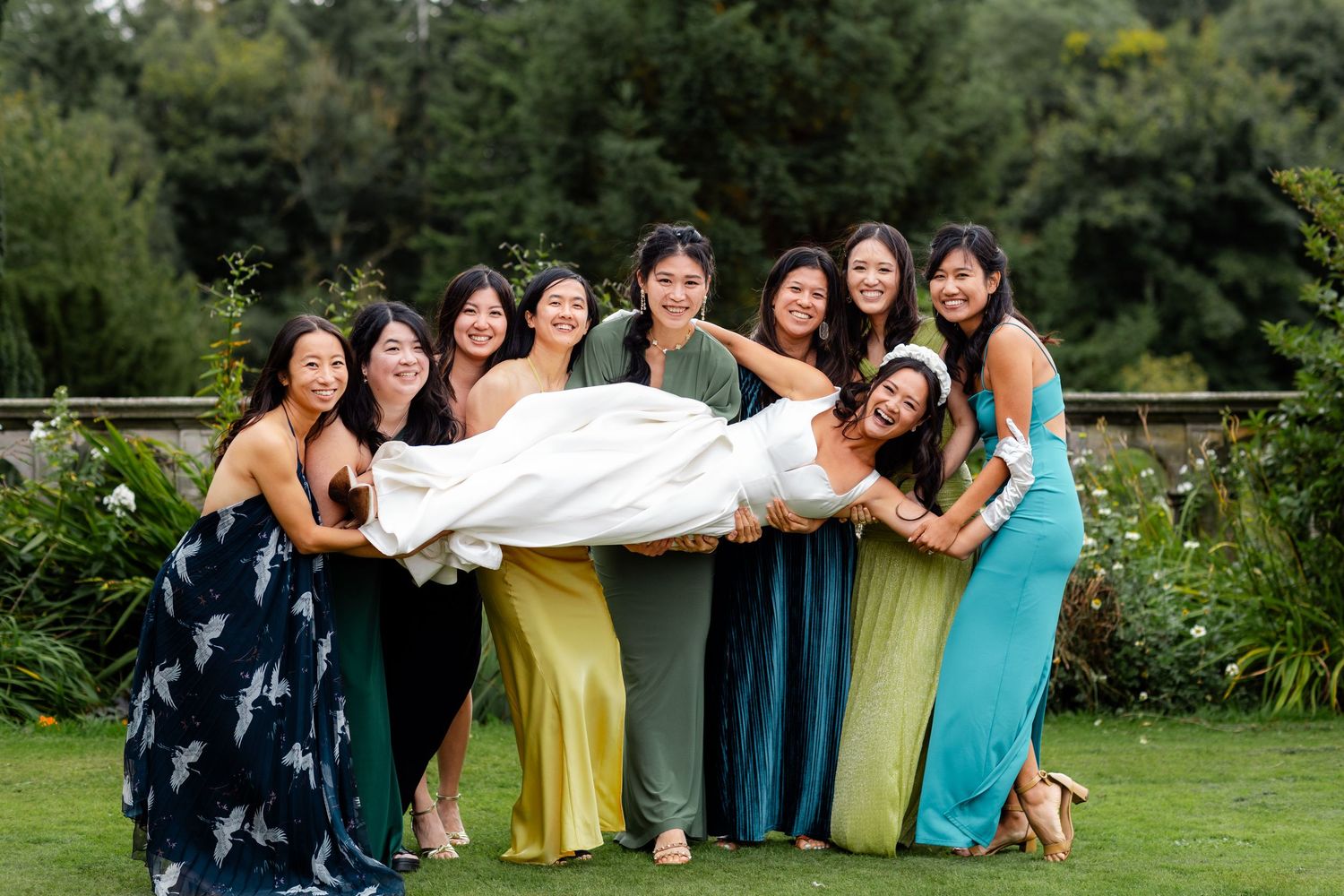 Bride in white gown lifted by smiling bridesmaids in colorful dresses on green lawn at wedding.