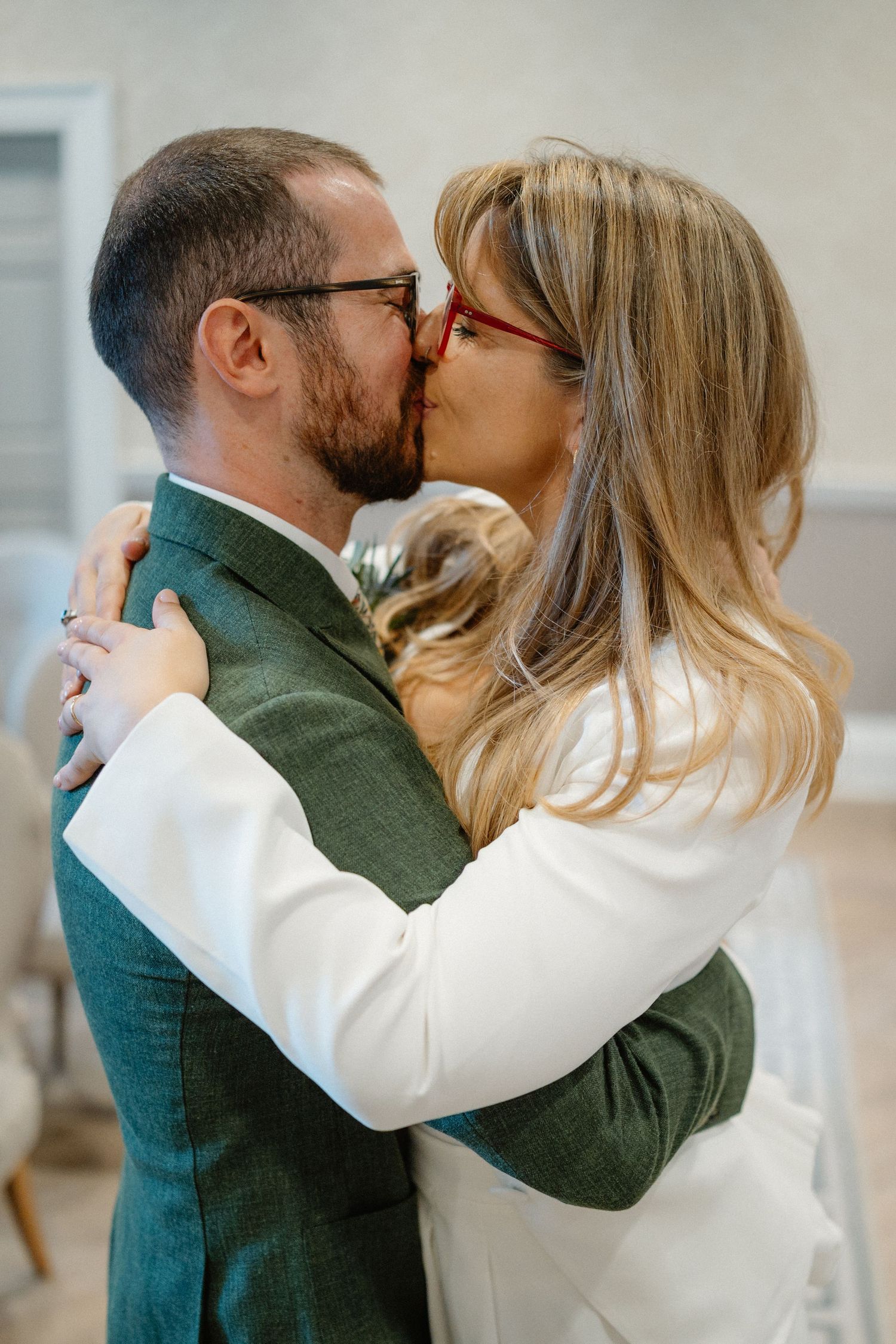 A couple sharing a romantic kiss, the bride in white and groom in a green suit, both wearing glasses.