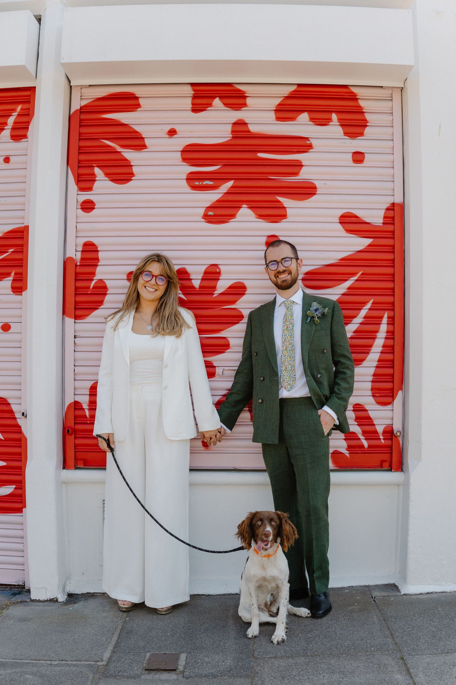 Newlywed couple in wedding attire holding hands with their spaniel dog in front of a red floral mural.
