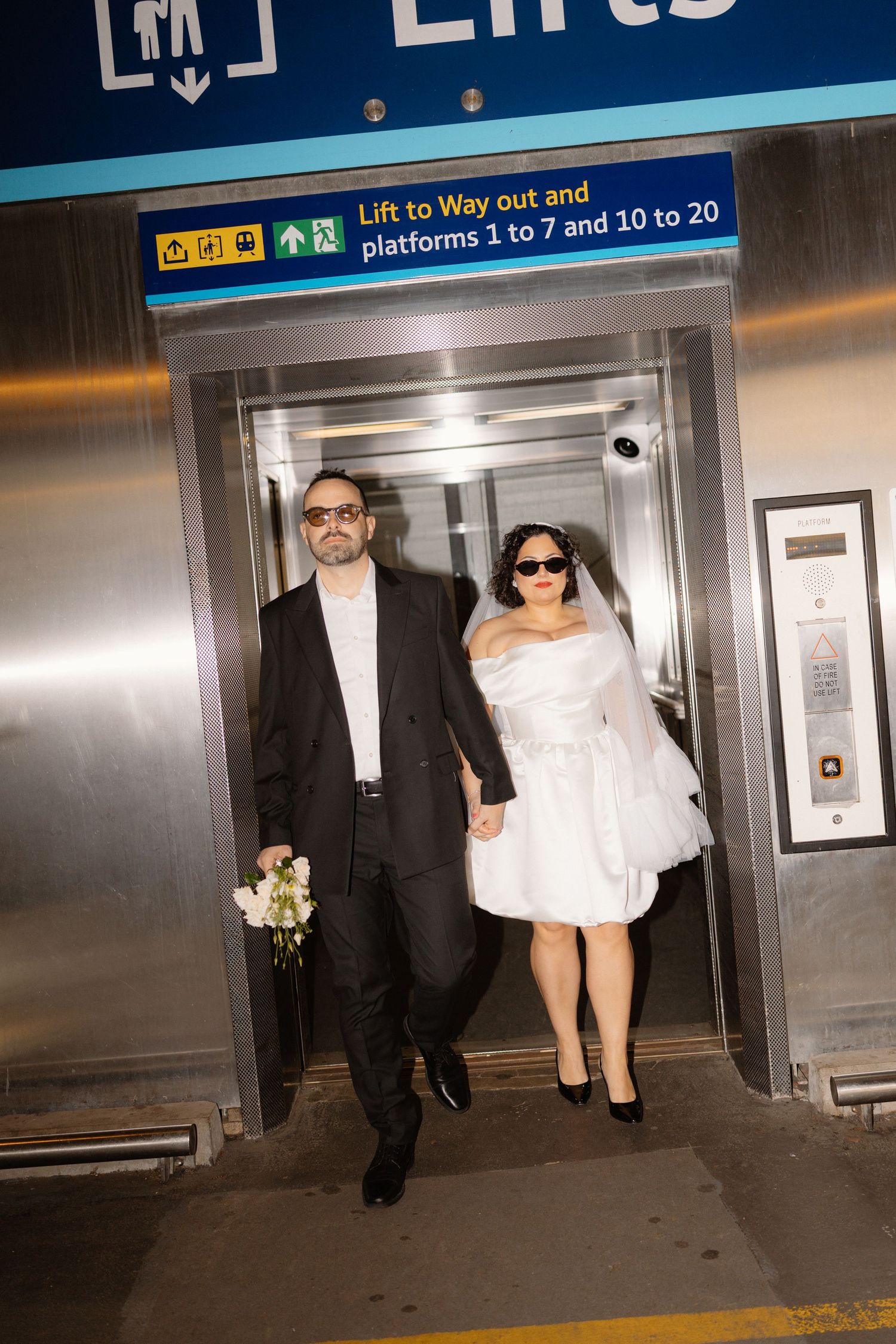 Bride and groom in sunglasses exit elevator at train station on their wedding day.