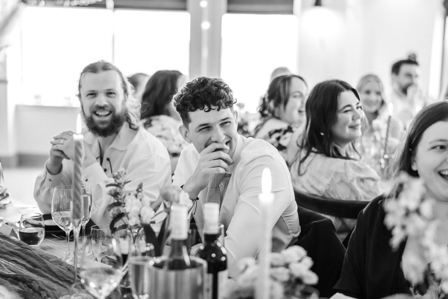Guests laughing and enjoying wedding reception dinner, black and white candid photography.