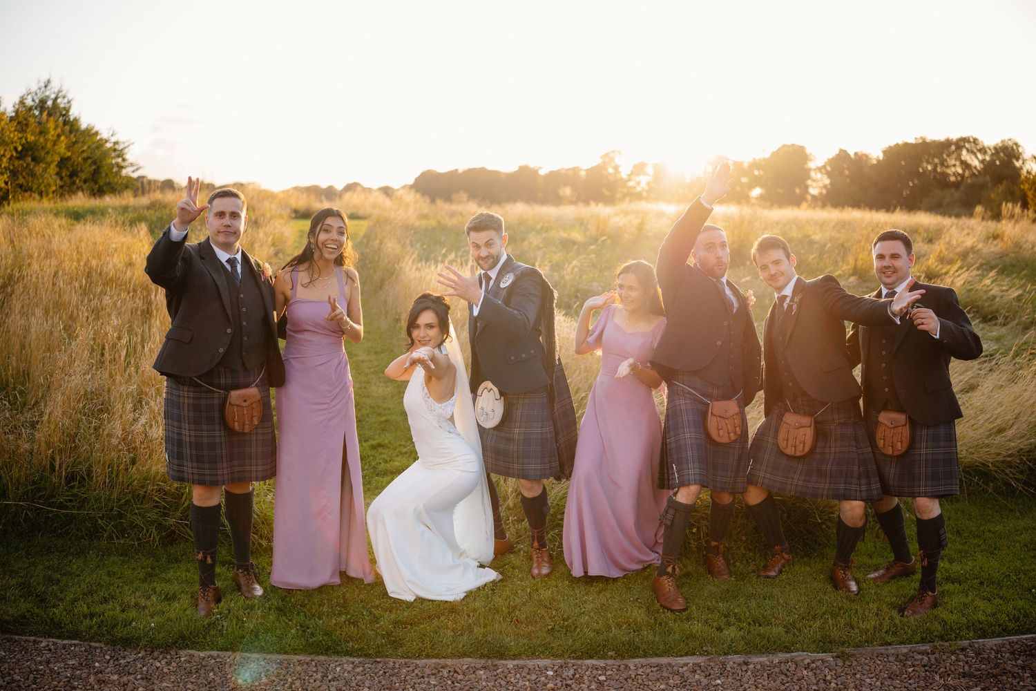 Wedding party in pink dresses and tartan kilts posing at golden hour in a scenic outdoor field.