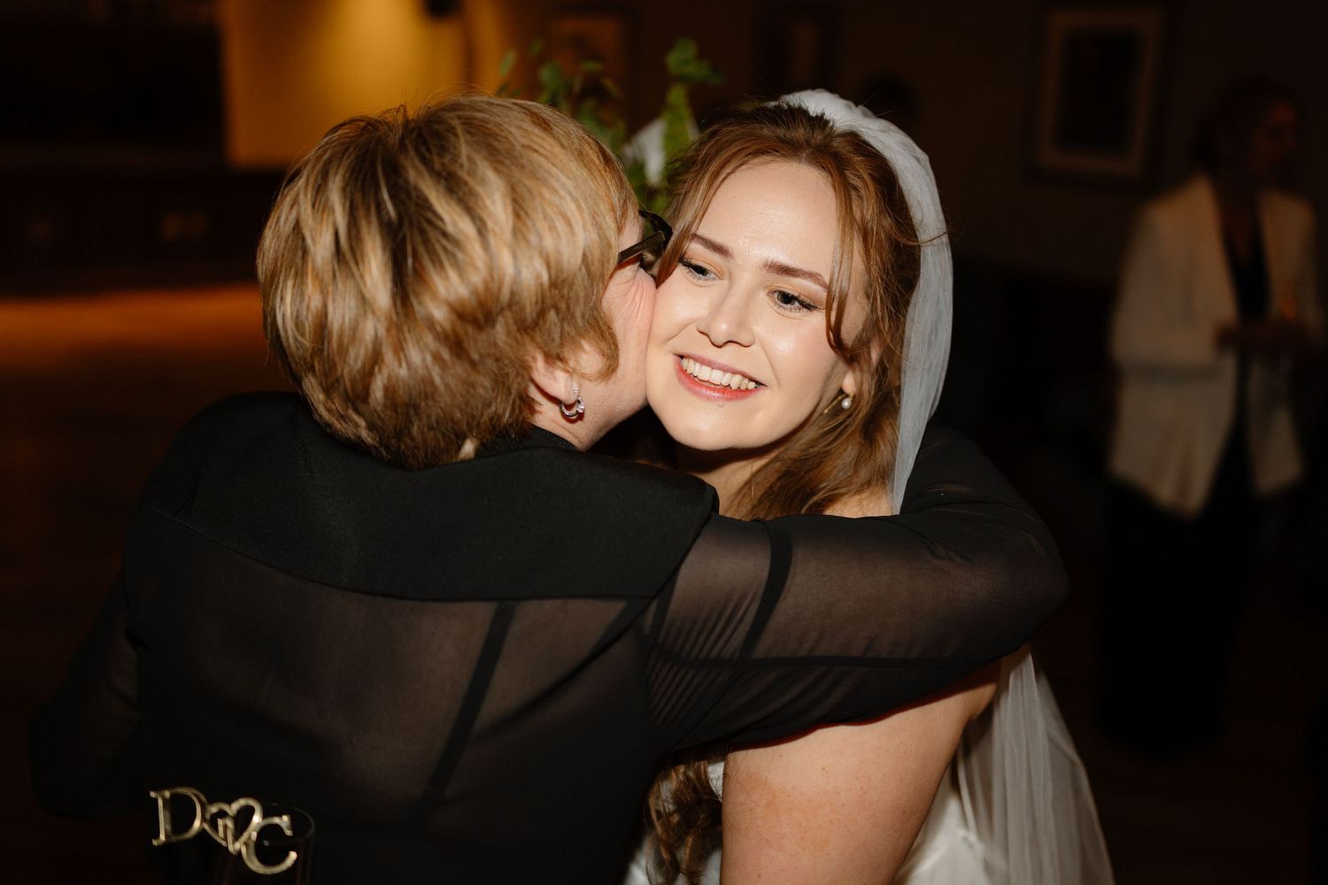 Bride in white veil smiling and hugging a woman in black dress at wedding reception.