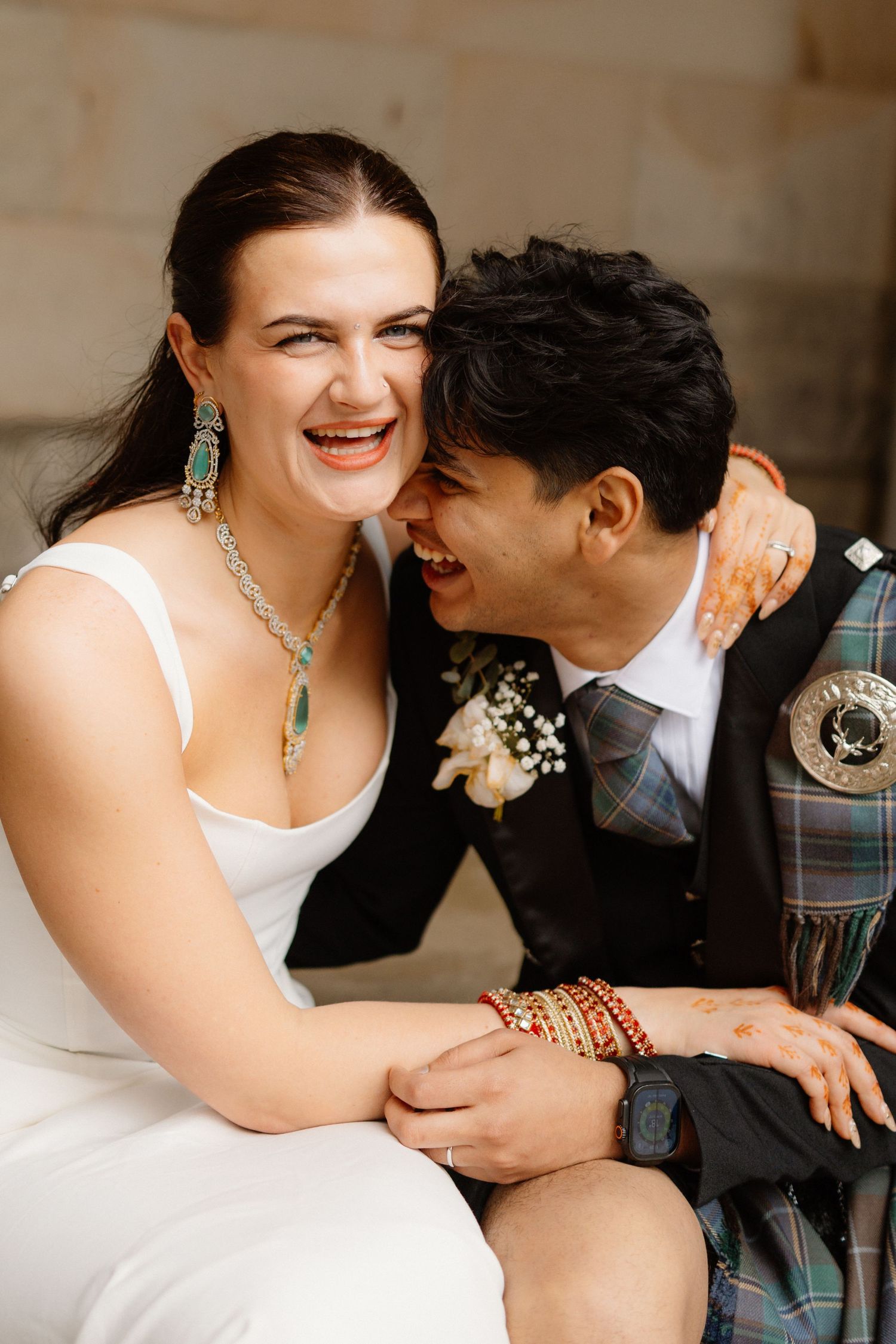 Joyful bride and groom laughing together, she in white dress with turquoise jewelry, he in traditional Scottish tartan kilt.
