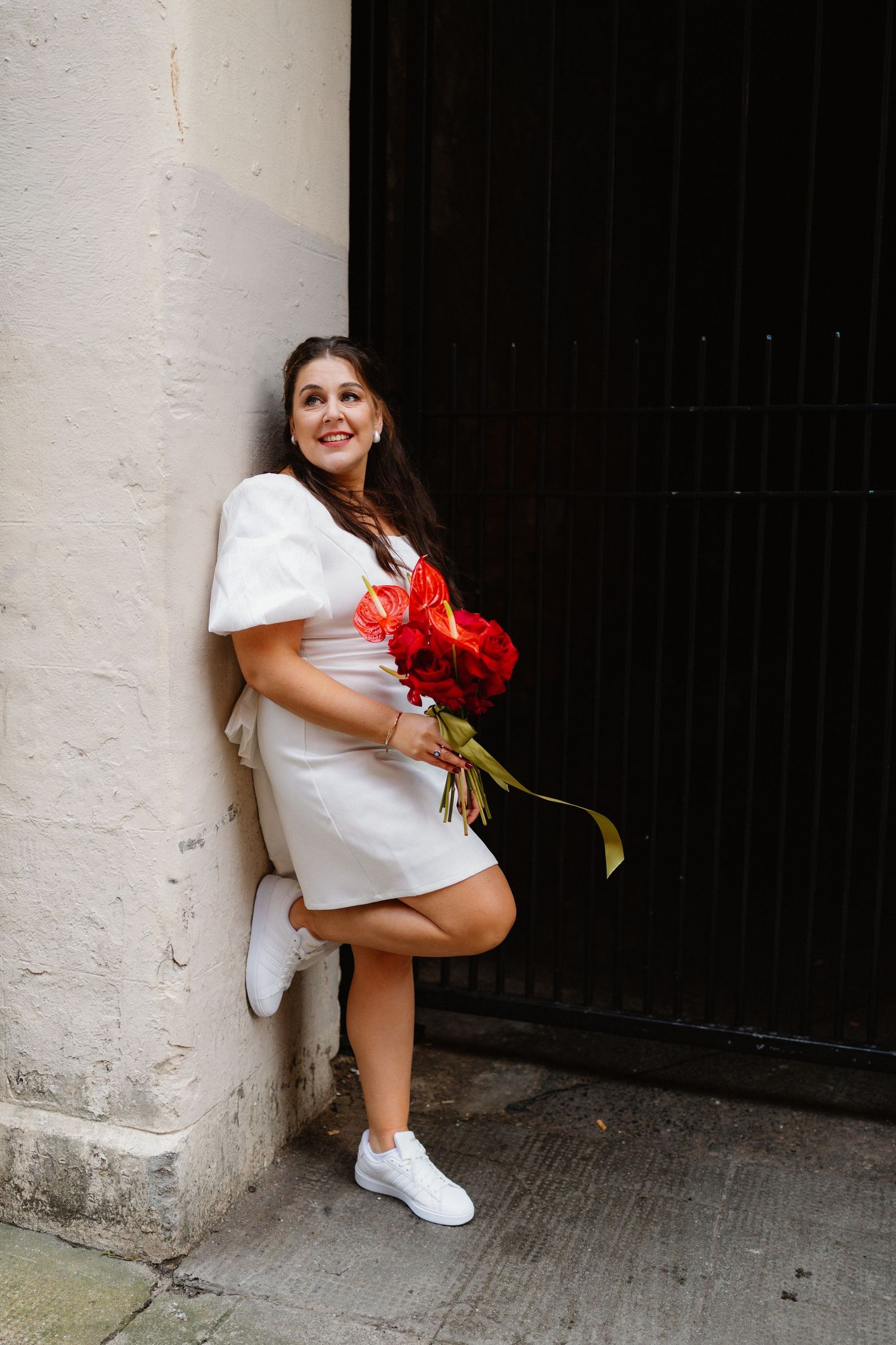 Woman in white dress holding red roses bouquet, leaning against wall outdoors, smiling, wearing white sneakers.
