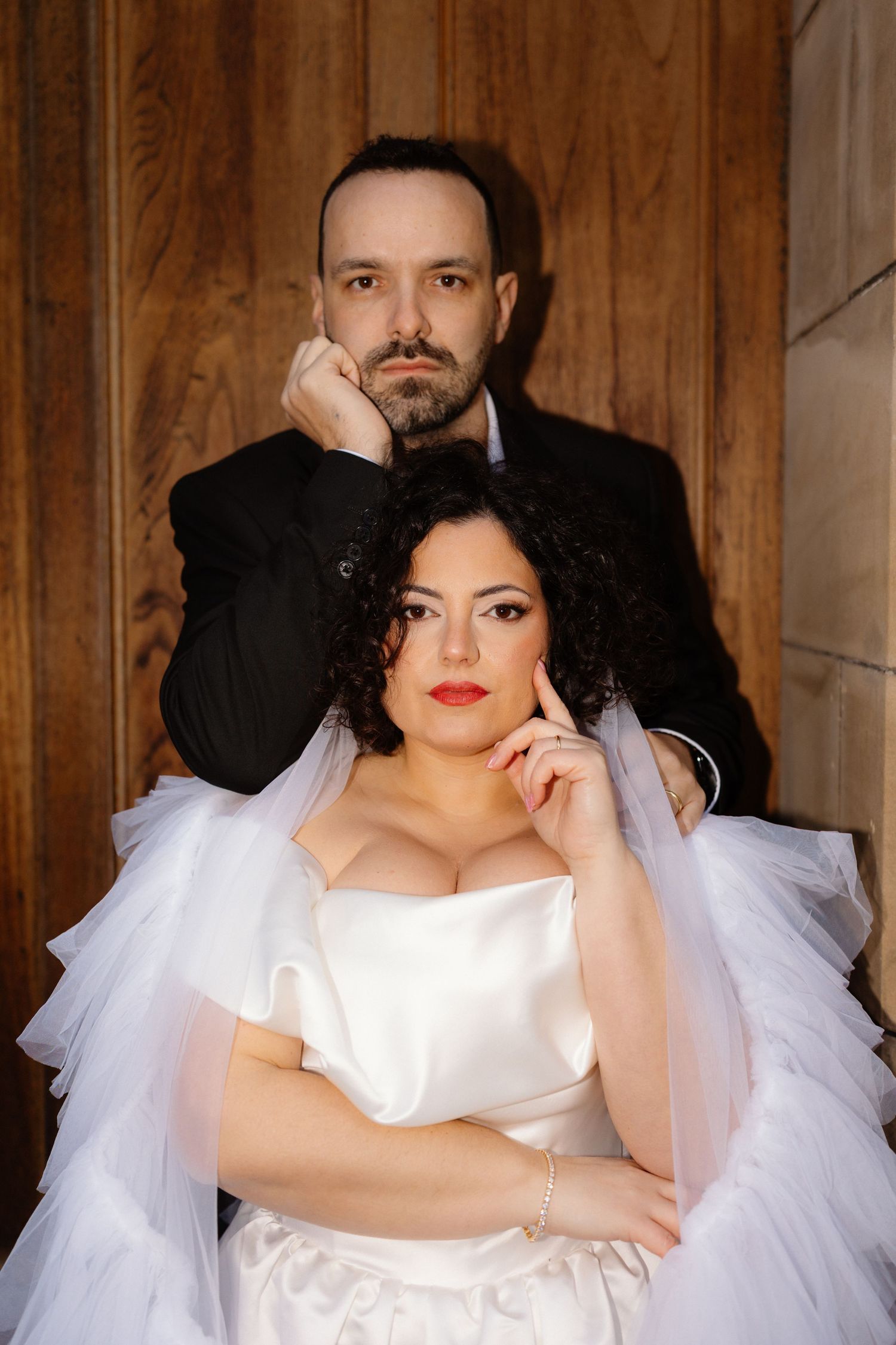 Elegant bride in white satin gown with veil and groom in black suit posing together for formal wedding portrait.