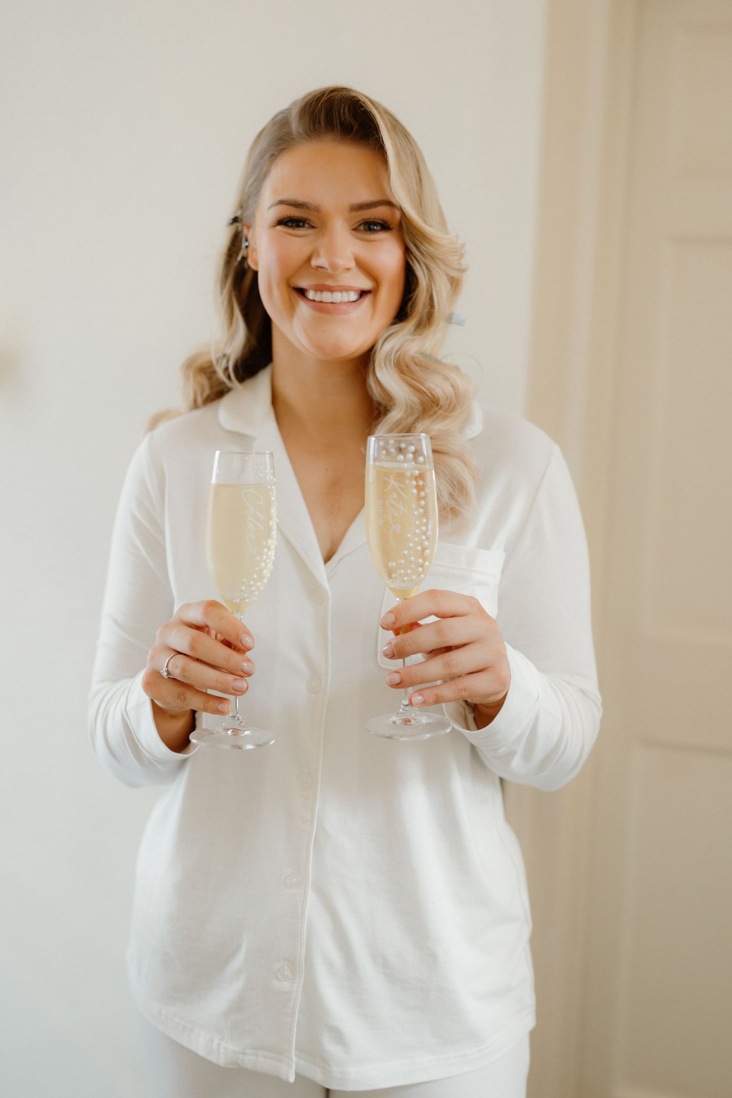 Smiling bride in white pajamas holding two champagne flutes, celebrating before her wedding ceremony.