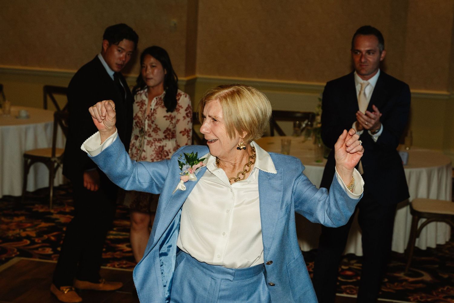 A joyful woman in a blue suit dances at a wedding reception while guests applaud in the background.