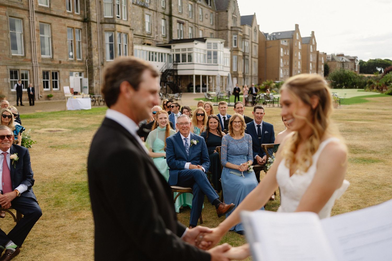 Bride and groom holding hands during an outdoor wedding ceremony with guests seated in front of a grand estate.