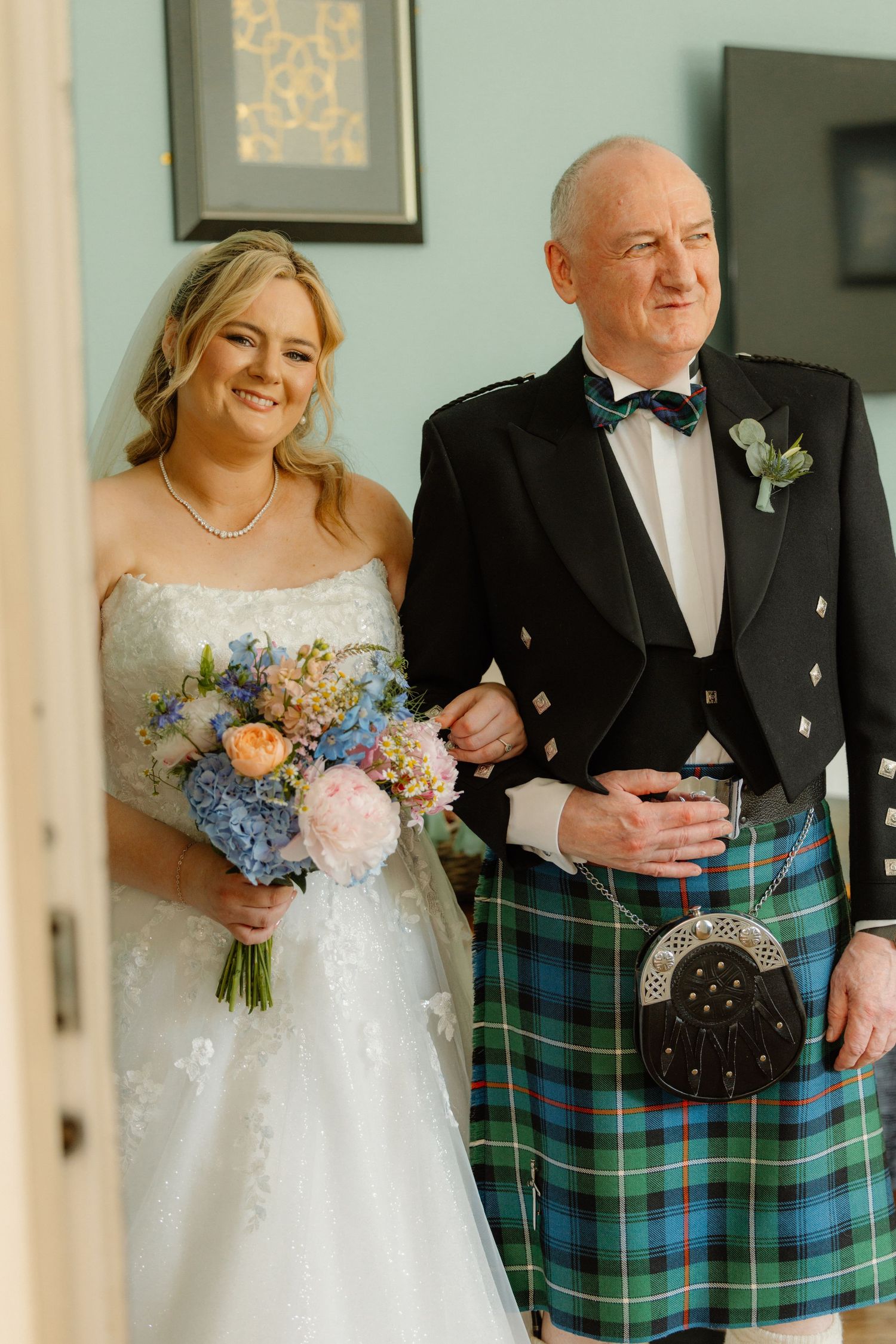 Smiling bride in strapless lace gown holding blue and pink bouquet with father in Scottish kilt.