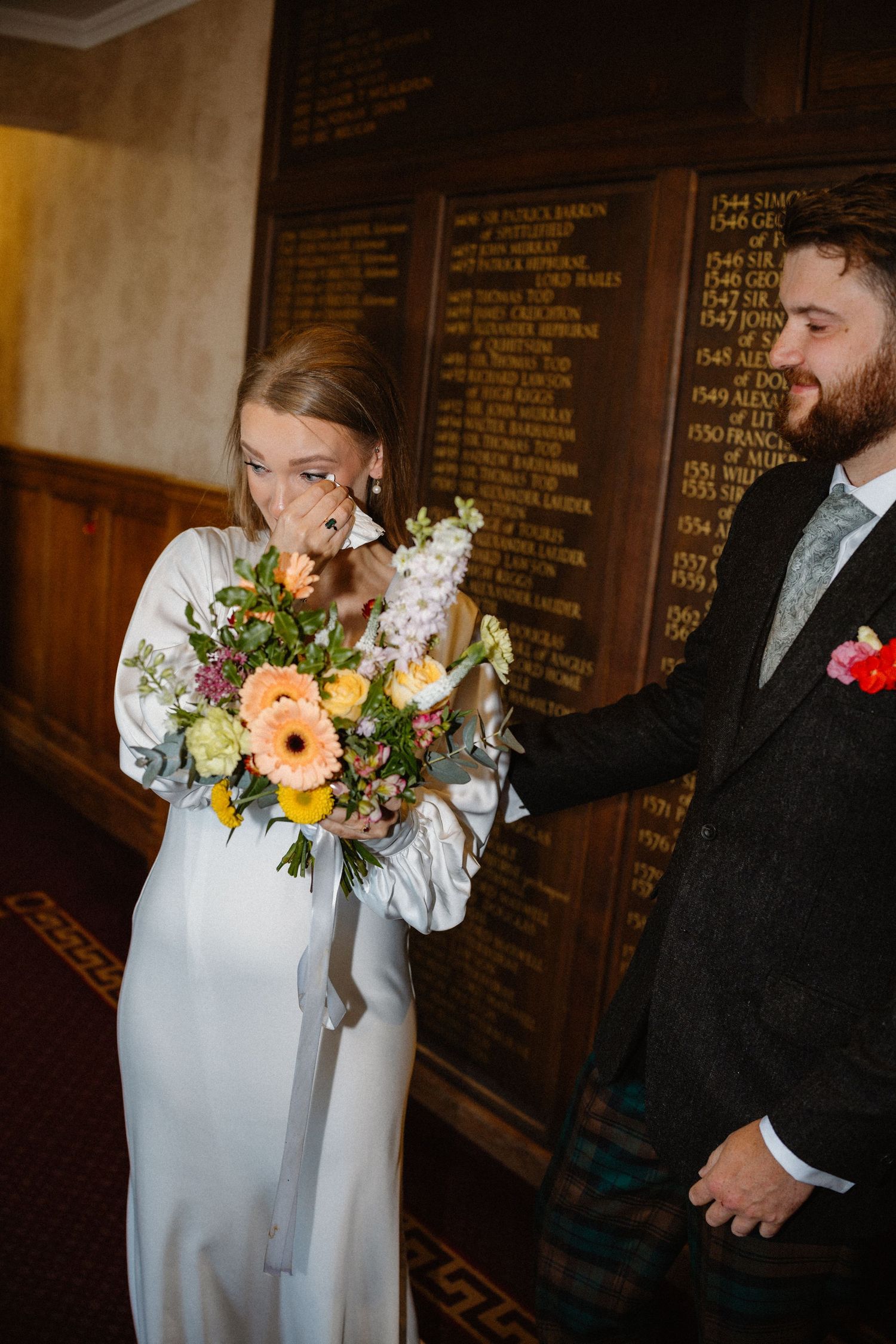 Emotional bride smelling colorful wedding bouquet beside groom in dark suit, standing before historic wooden honor board.