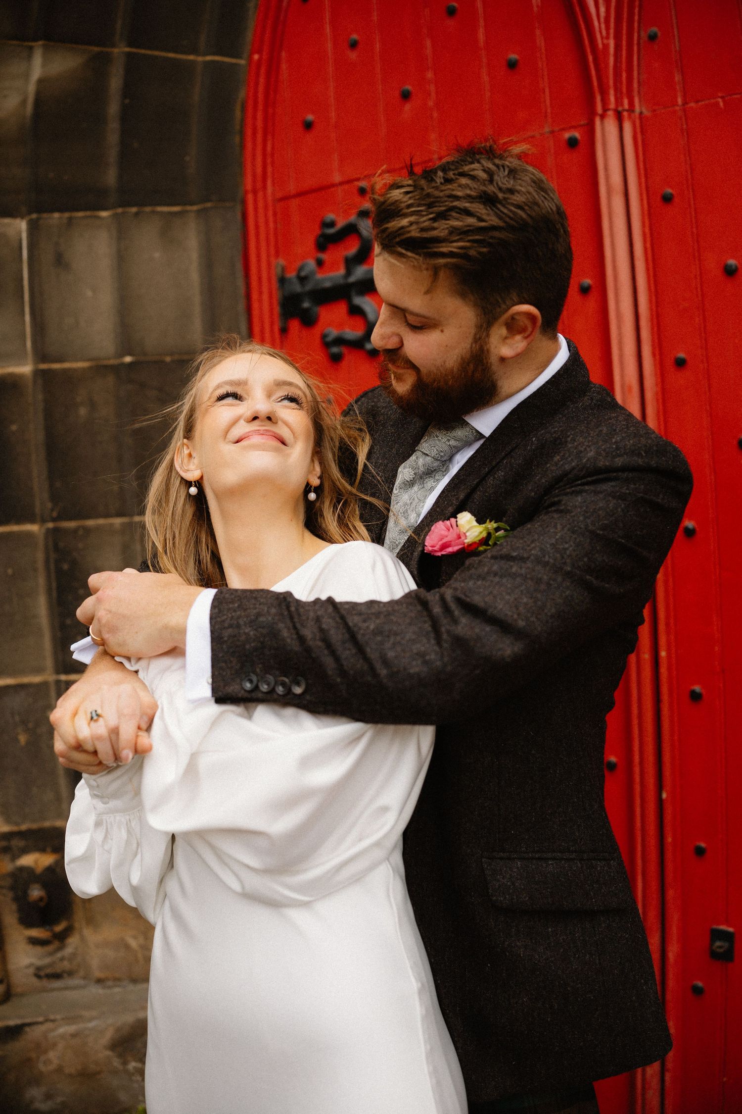 Groom embraces smiling bride in white dress before a vibrant red church door.