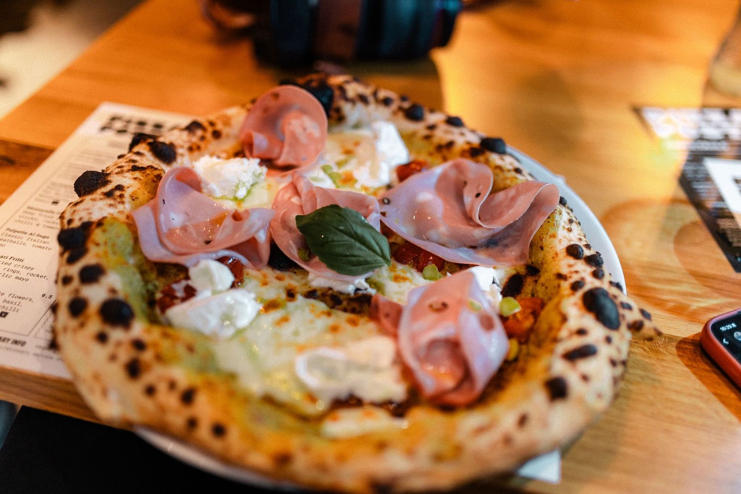 Authentic Neapolitan pizza topped with mortadella, mozzarella and basil on wooden restaurant table.