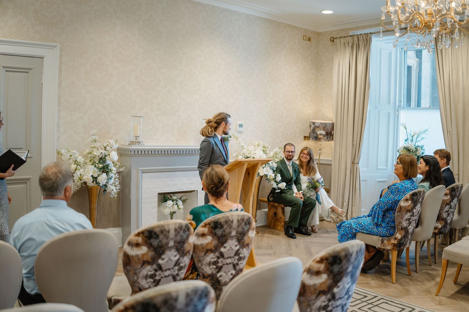 Intimate wedding ceremony in an elegant room with floral arrangements, guests seated in patterned chairs facing the couple.