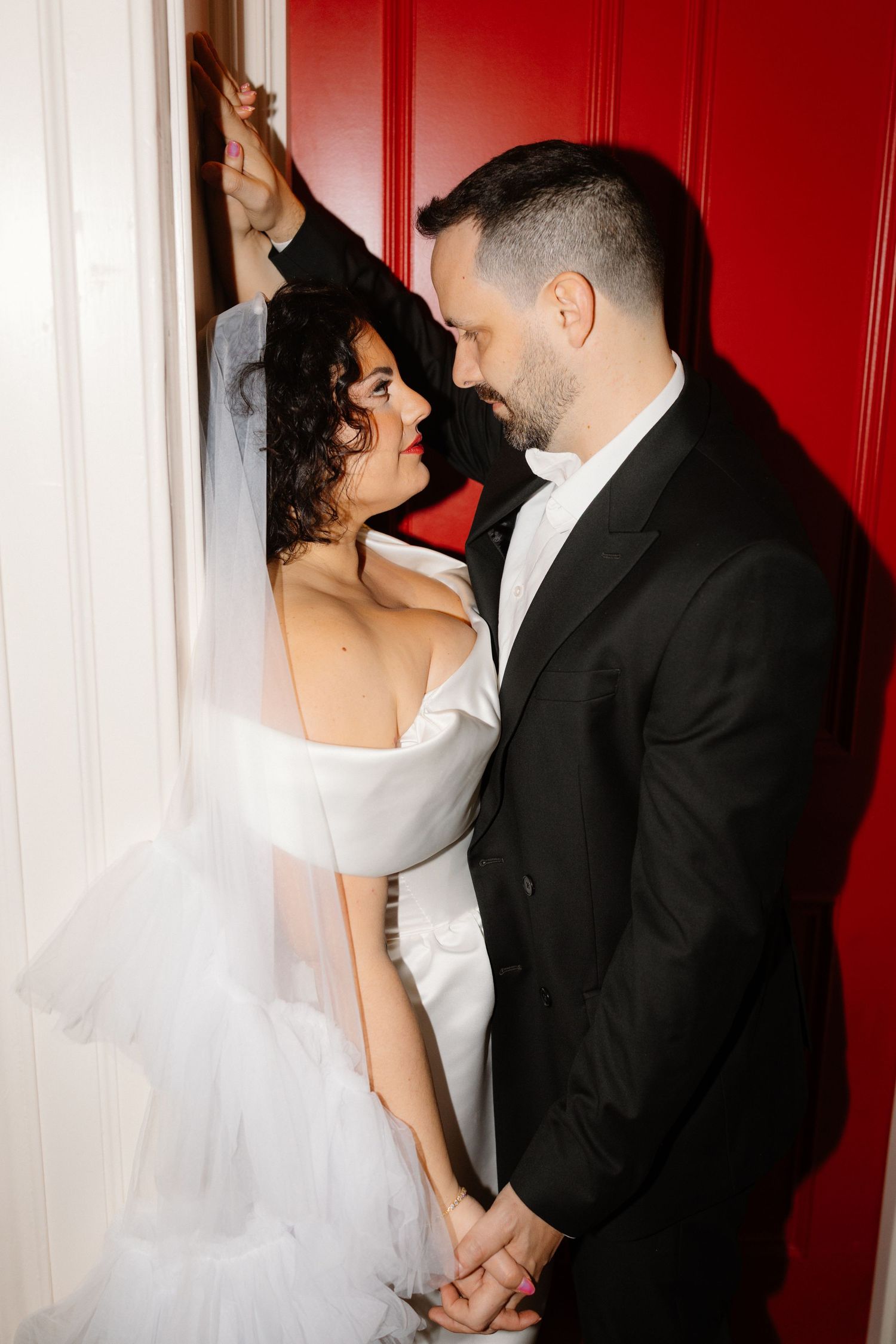 A bride in a white off-shoulder gown and groom in black tuxedo share a romantic moment against a red door.