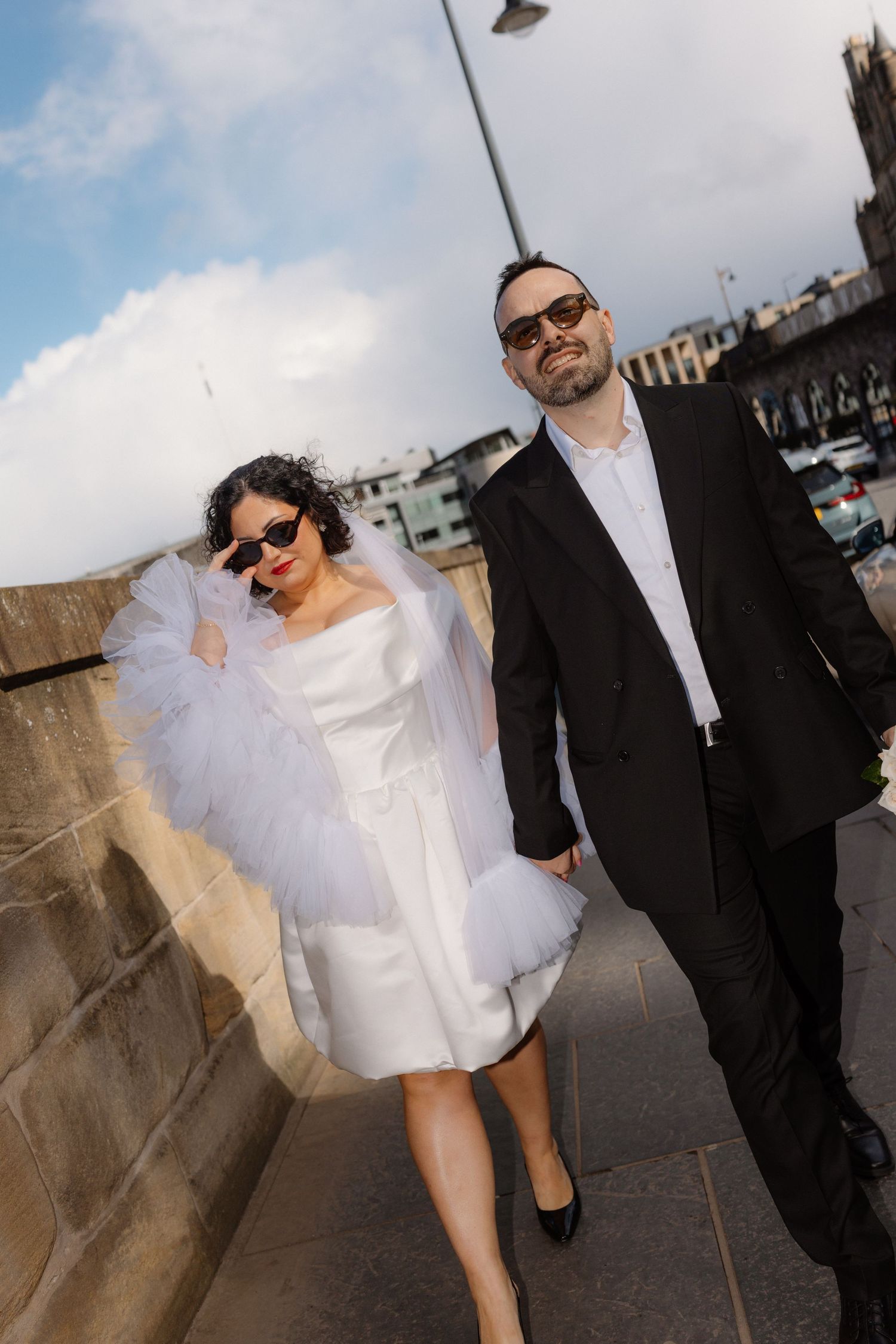 Stylish bride in white mini dress with tulle wrap and groom in black suit walking together in Edinburgh.
