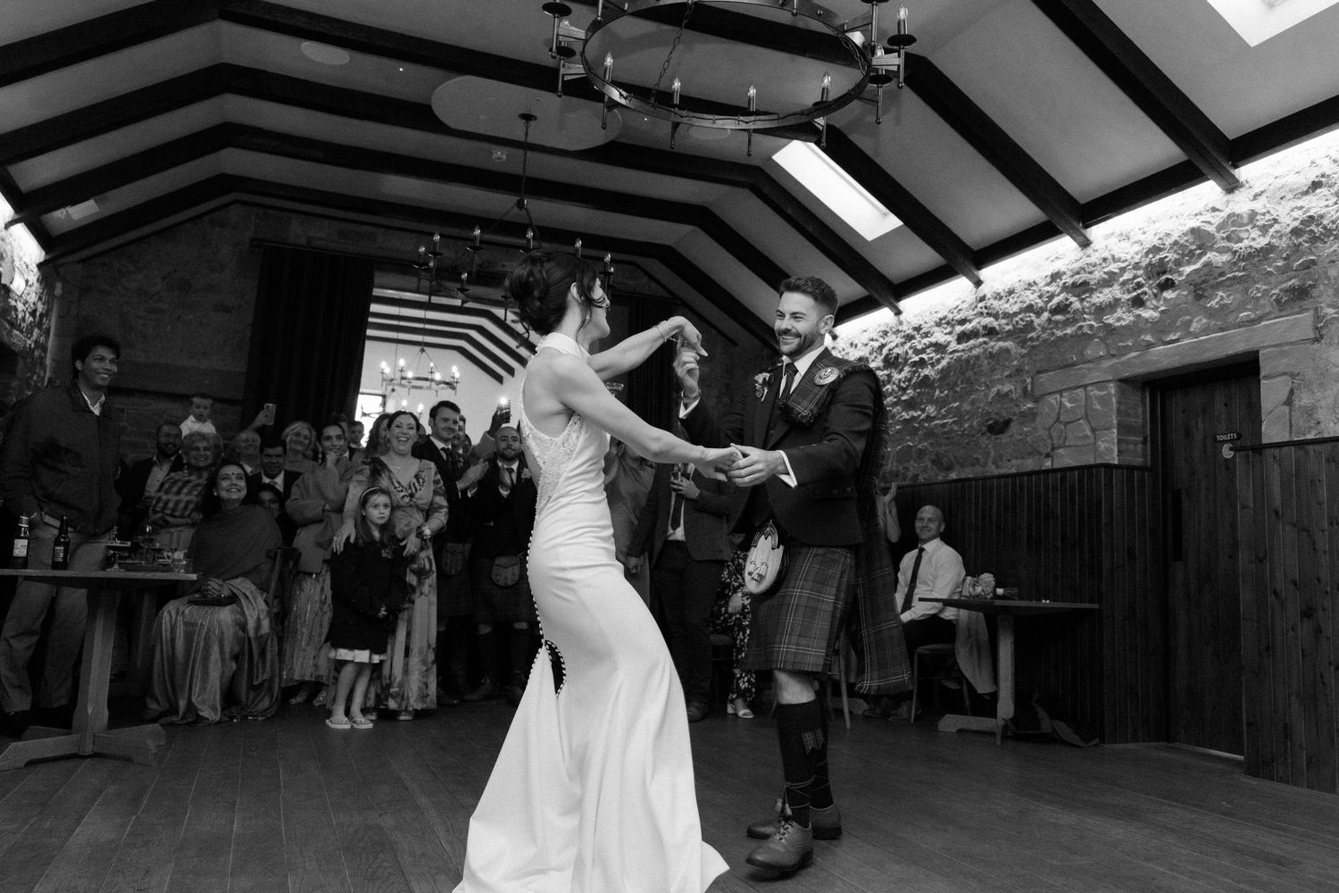 Bride and groom share their first dance in a rustic wedding venue while guests watch and celebrate.