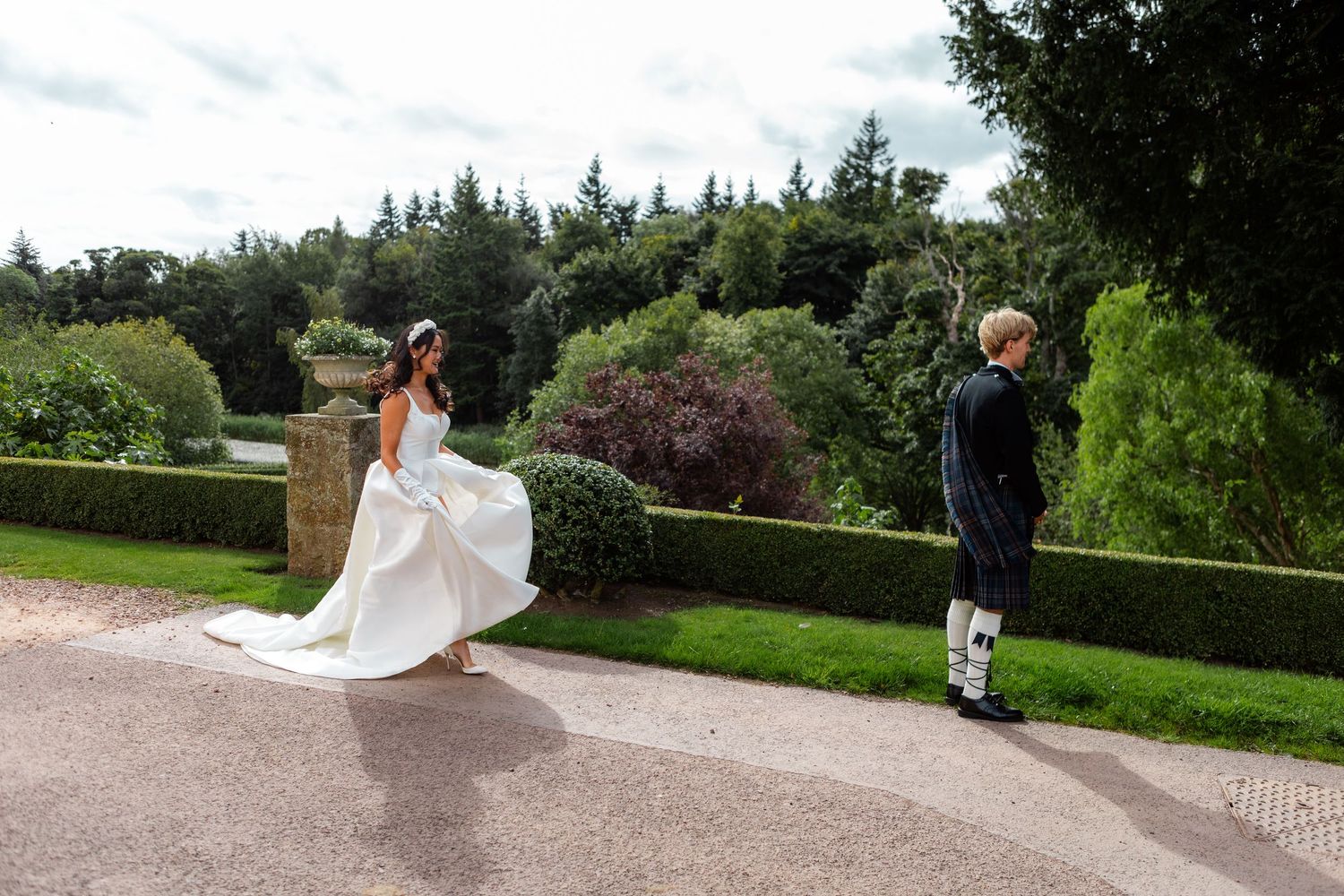 Bride in white gown twirls on garden path as groom in Scottish kilt stands nearby at outdoor wedding.