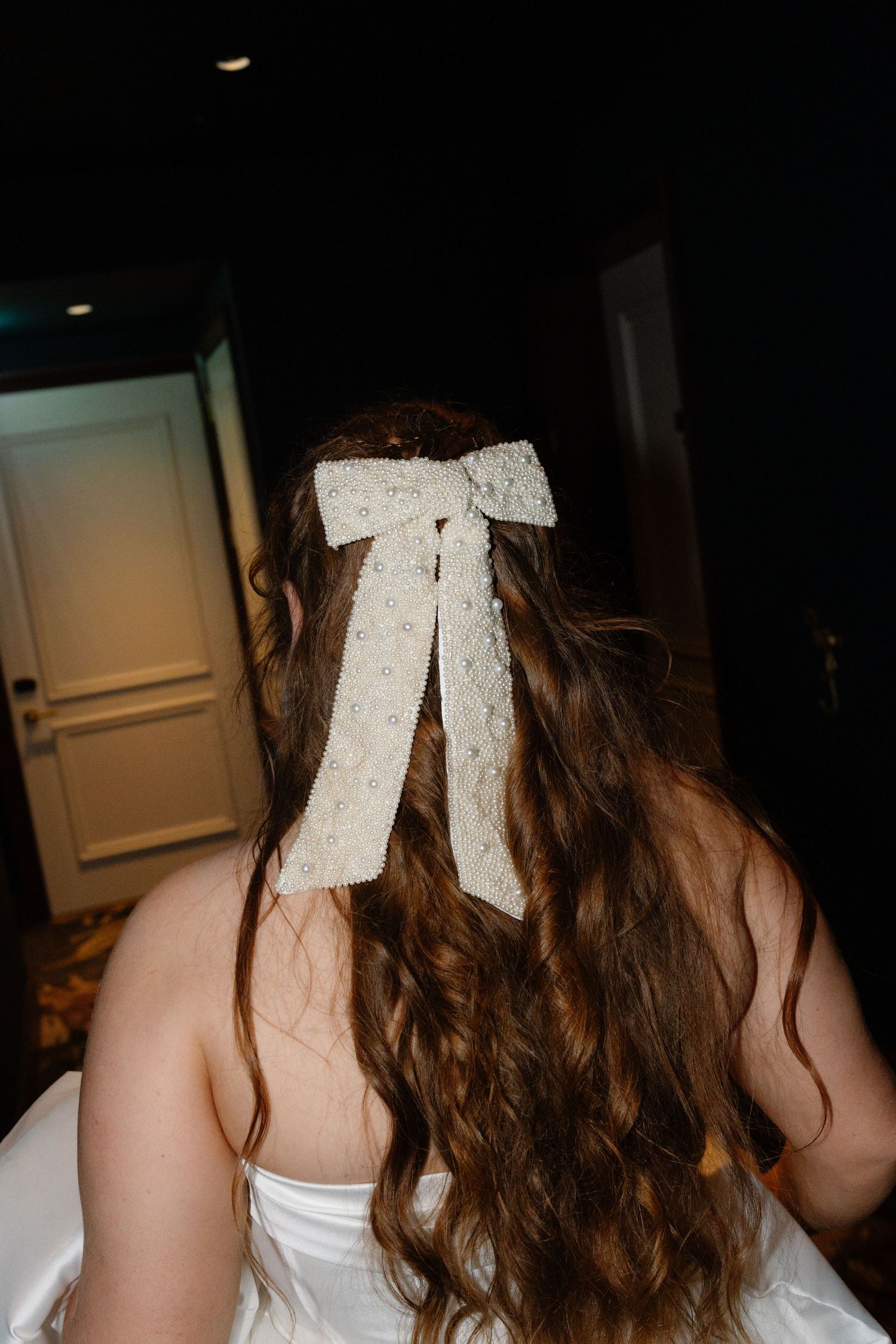 Bride with long wavy brown hair adorned with an elegant white lace bow hair accessory viewed from behind.