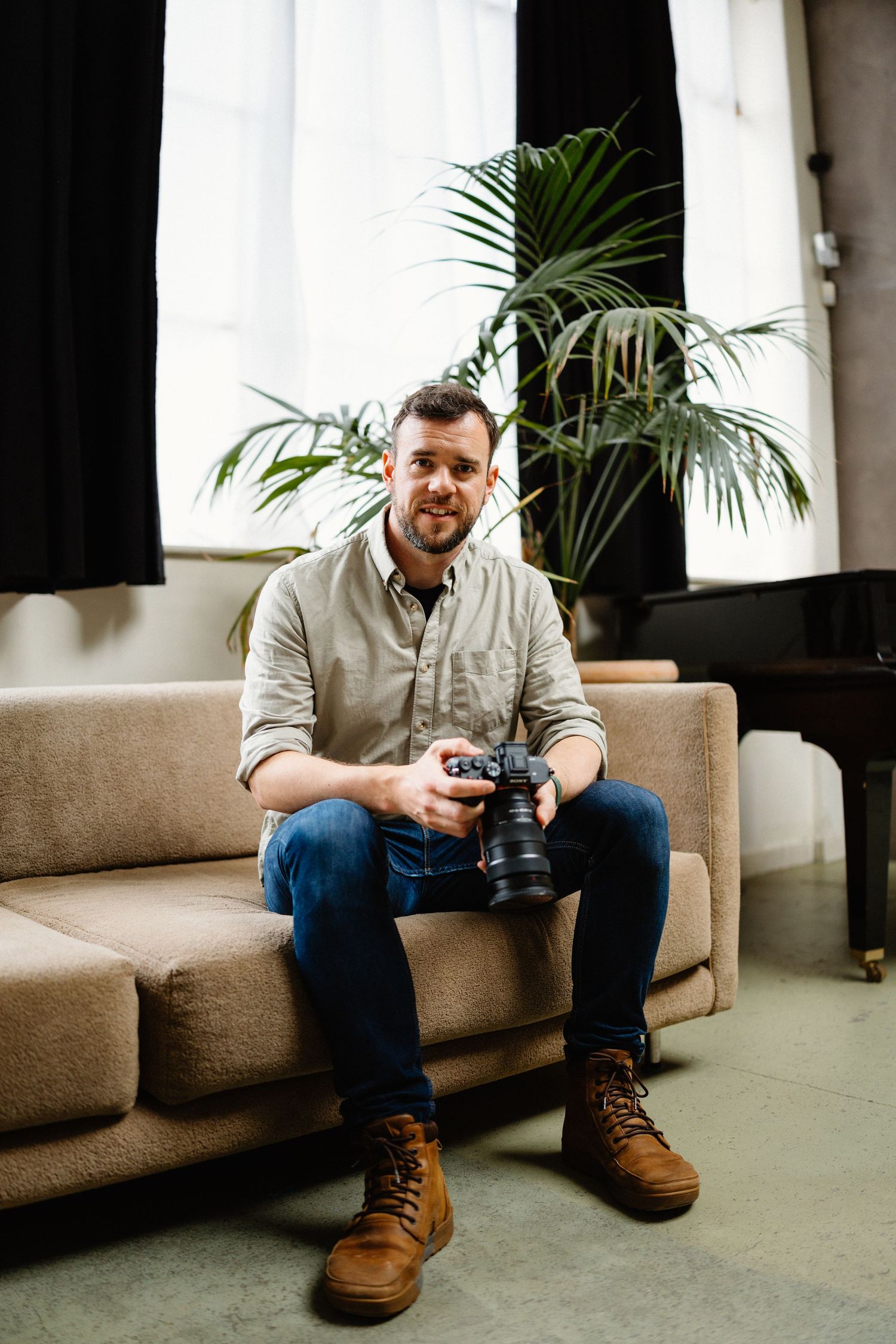 Photographer sits on brown couch with camera near indoor palm plant.