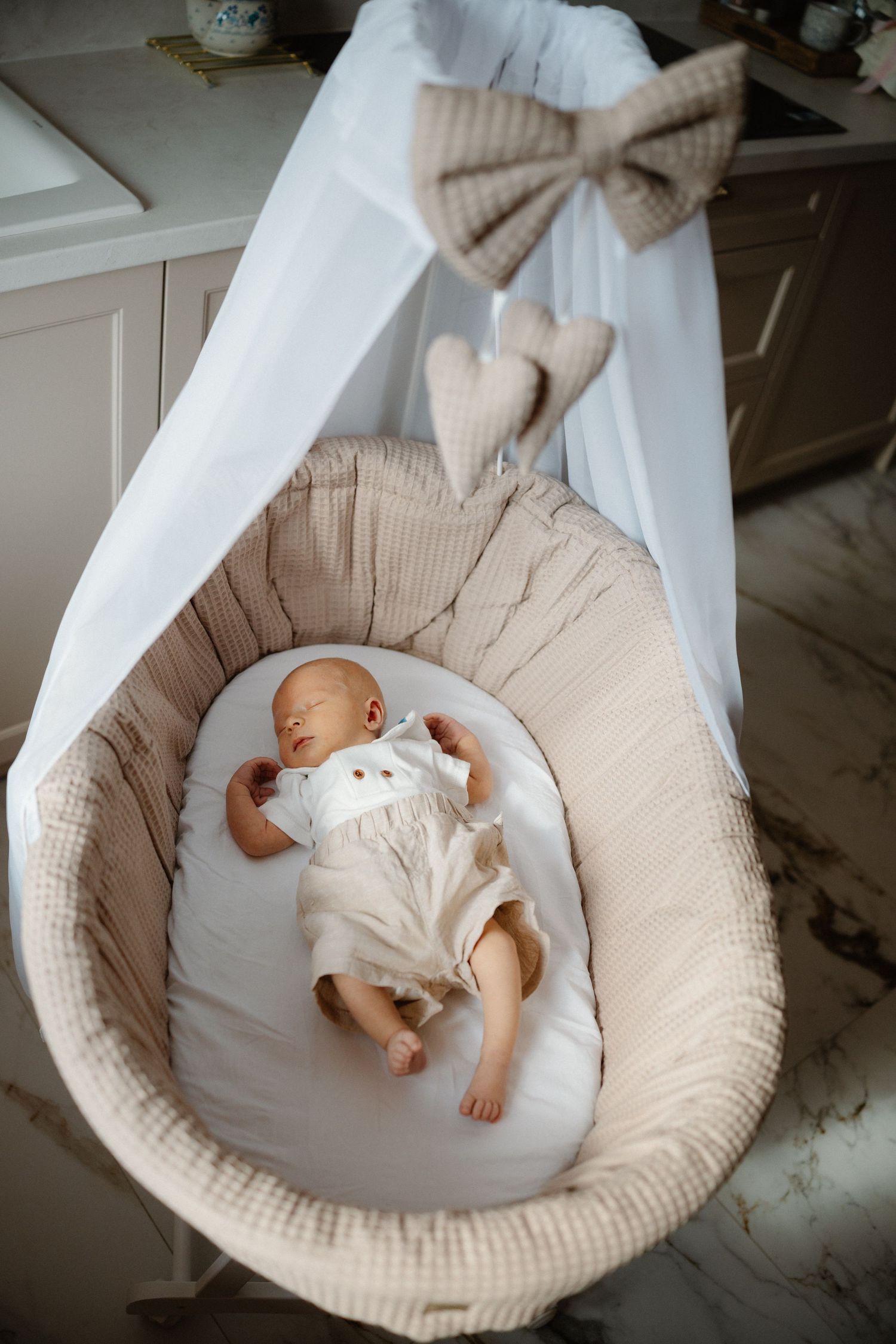 Newborn sleeping peacefully in beige circular bassinet with decorative bow.