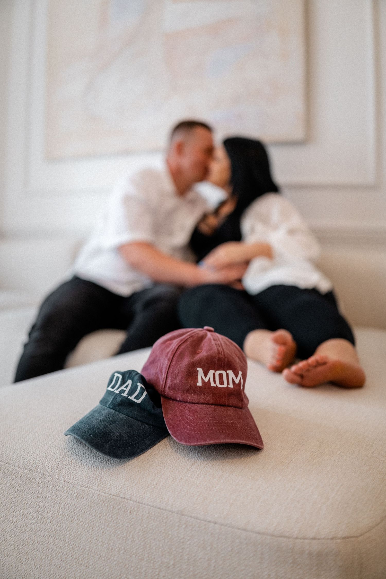 Two baseball caps, one burgundy with 'MOM' text and one black, displayed on a light colored couch.