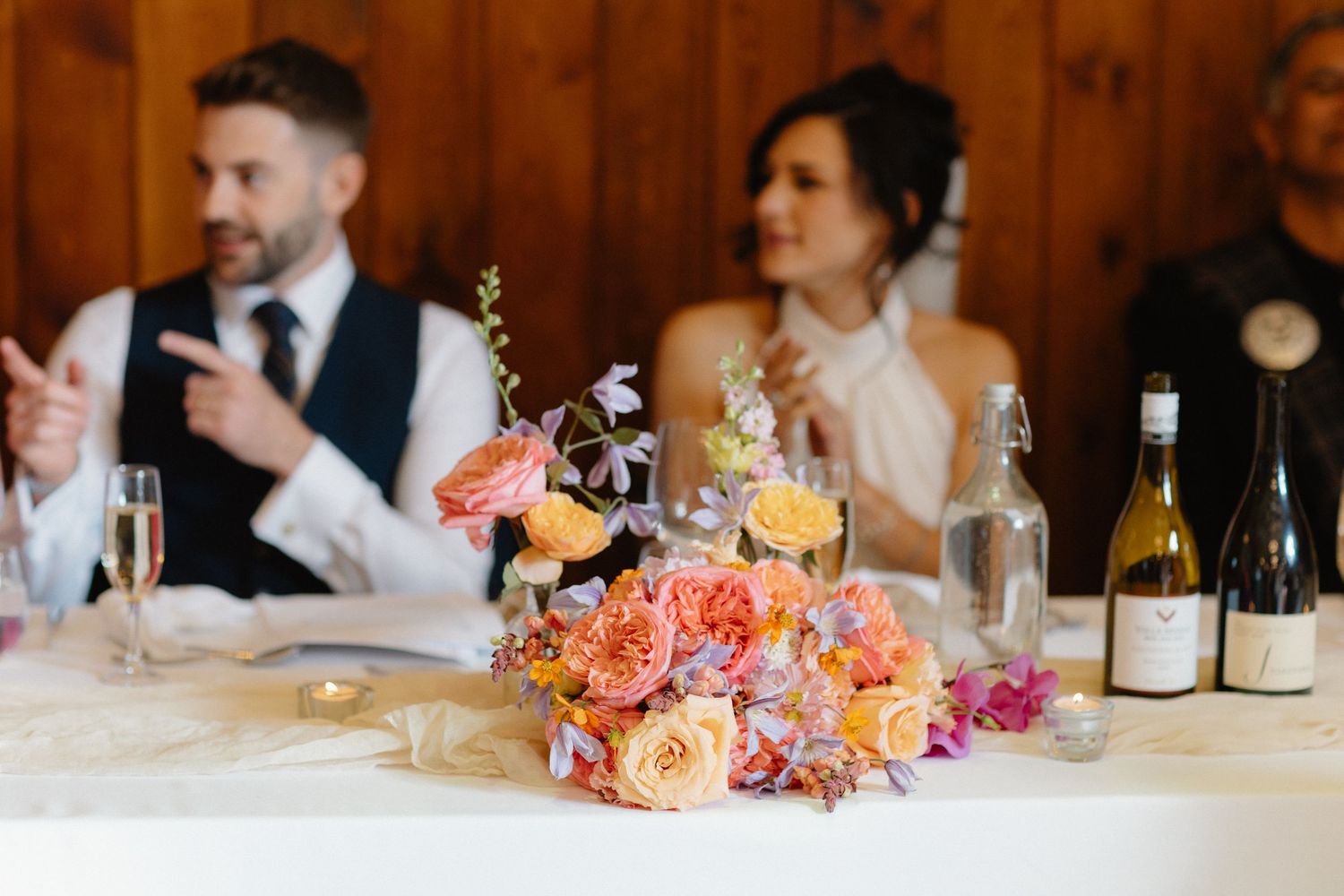 Wedding reception head table decorated with colorful floral arrangements and wine bottles.