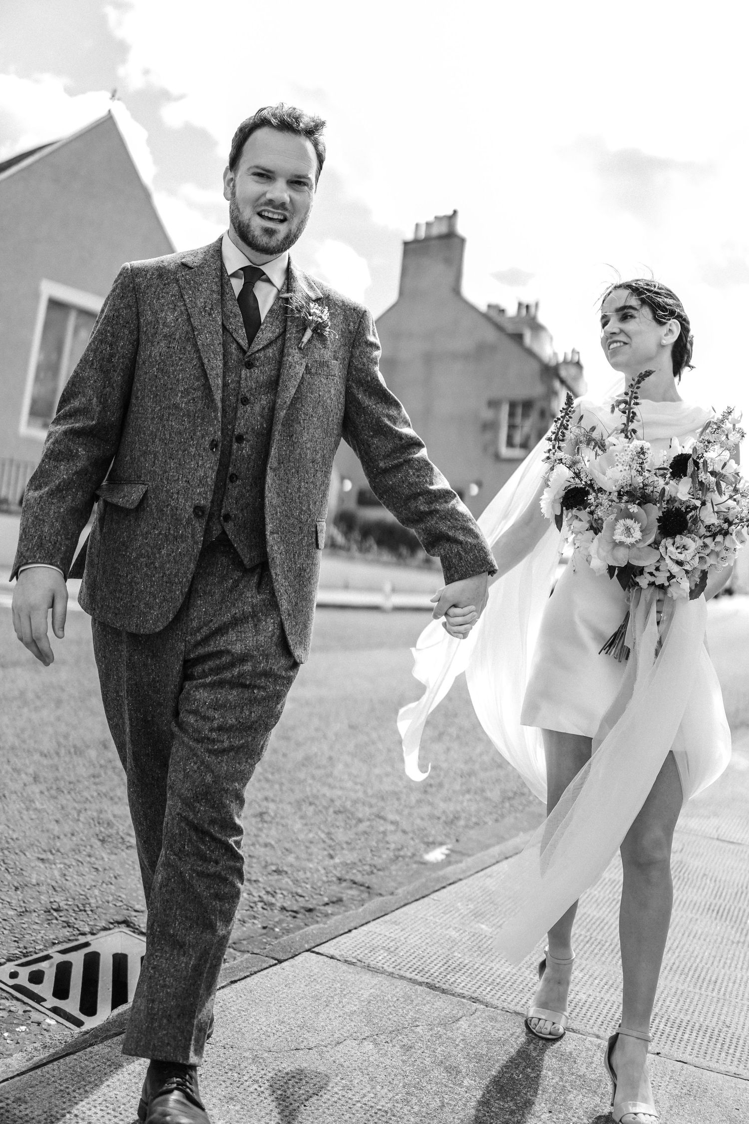 Wedding couple runs playfully through an urban setting in formal attire.