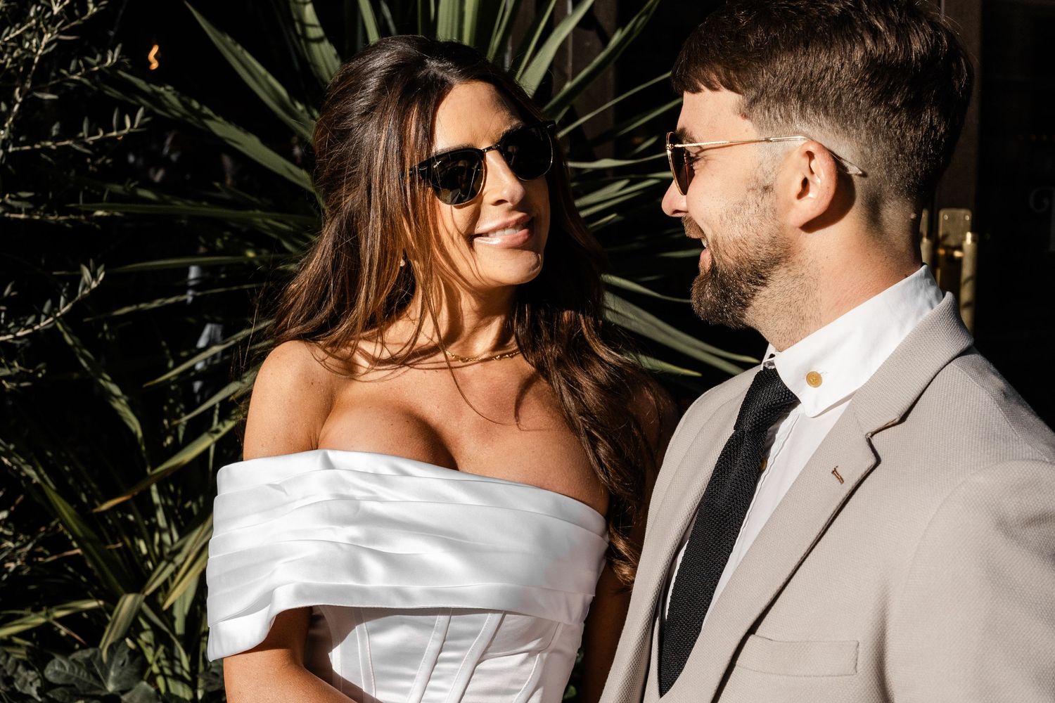 Stylish couple in sunglasses share a joyful moment against tropical foliage.