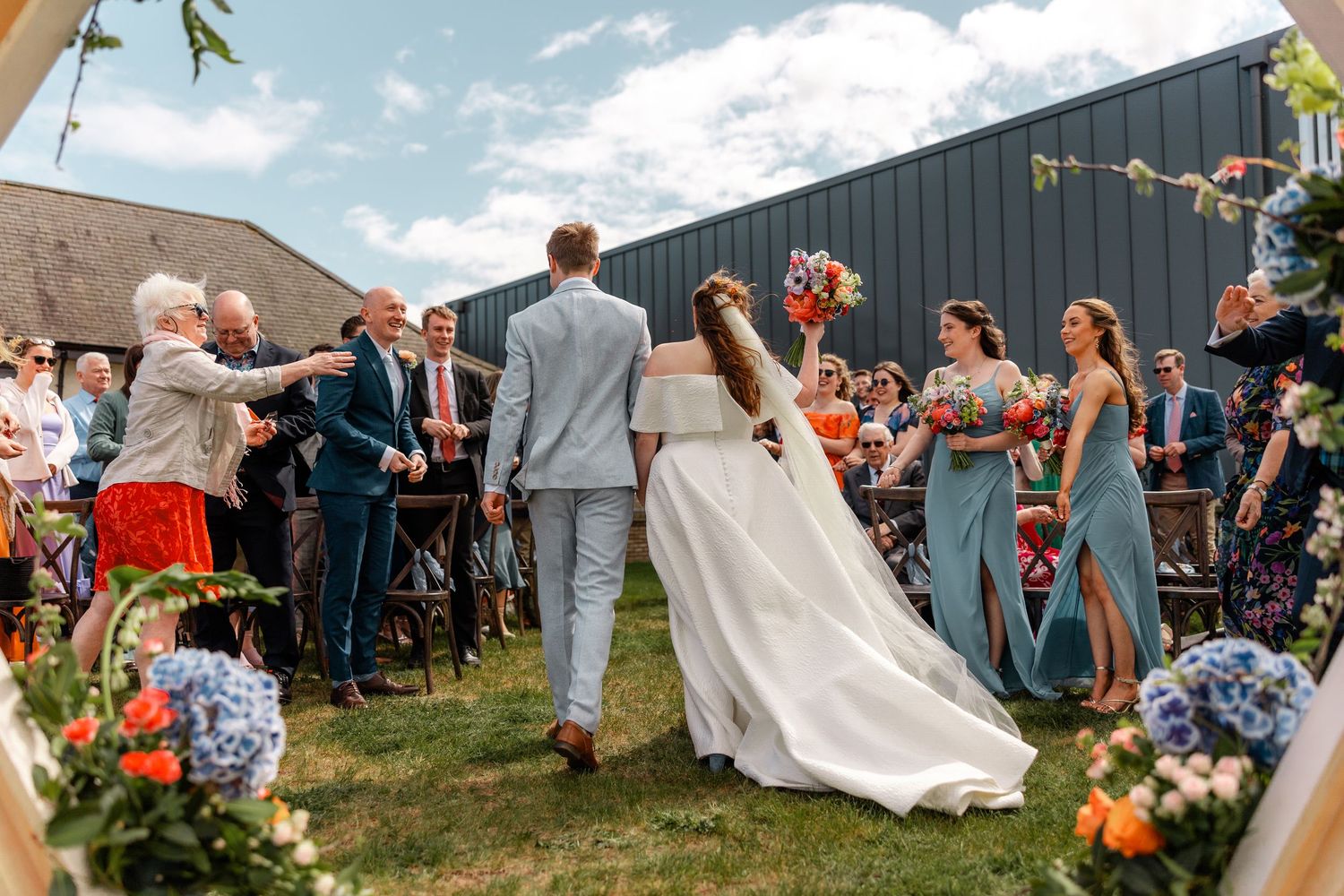Outdoor wedding ceremony with guests gathered under blue skies and decorative floral arrangements.