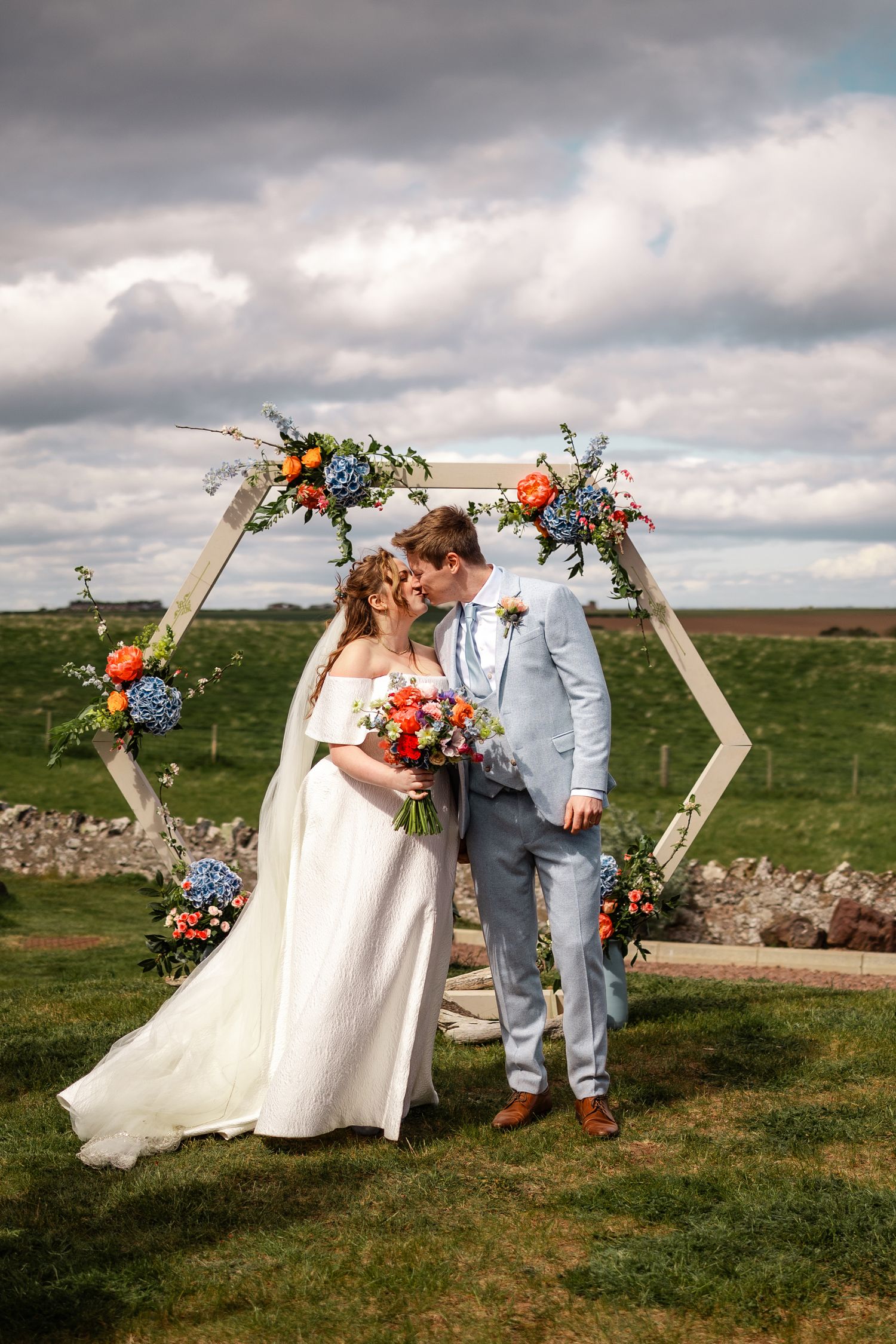 Couple poses under hexagonal floral arch with colorful bouquet in countryside setting.