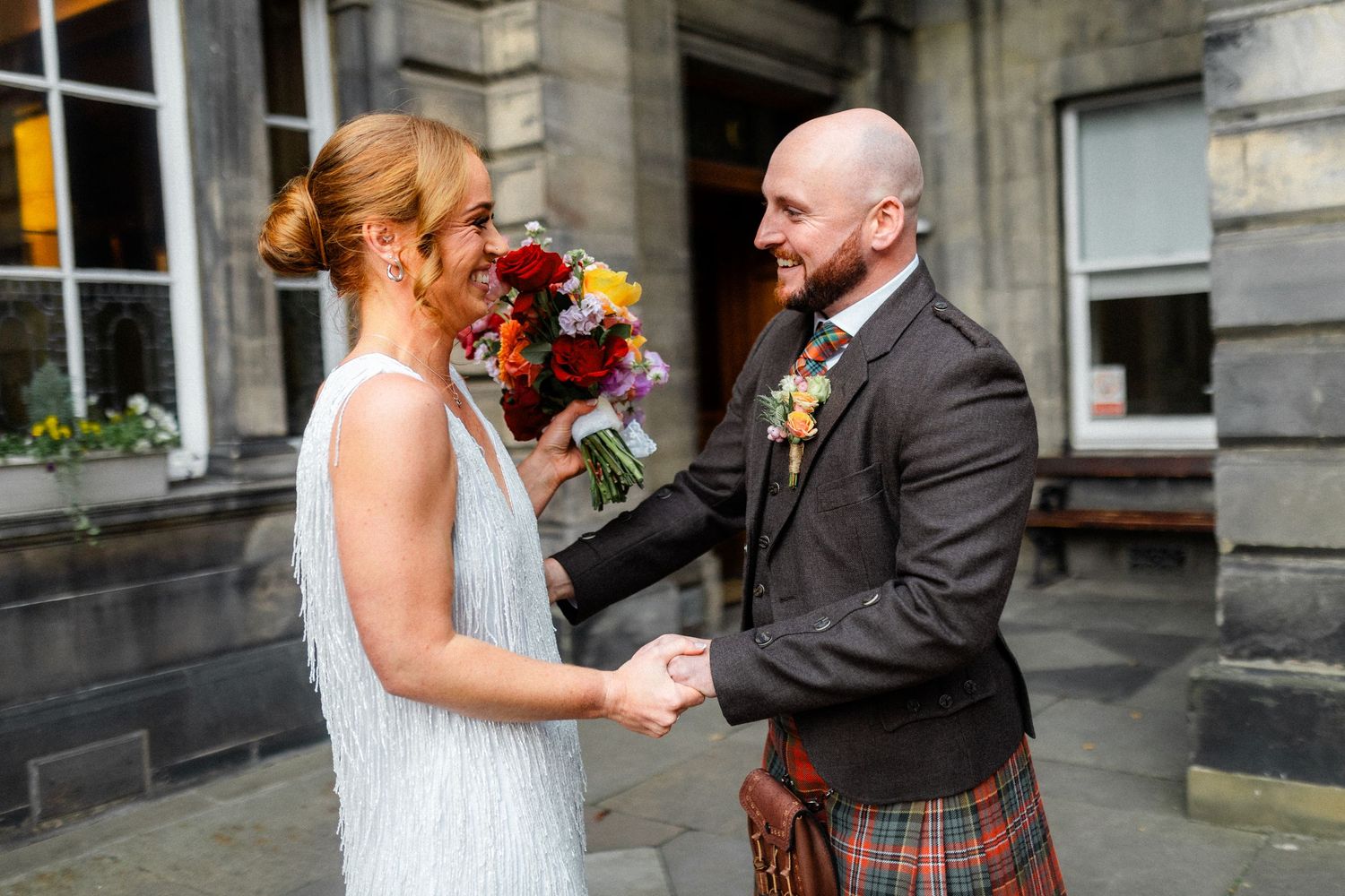 Newlyweds laughing together outside historic venue with bright floral bouquet.