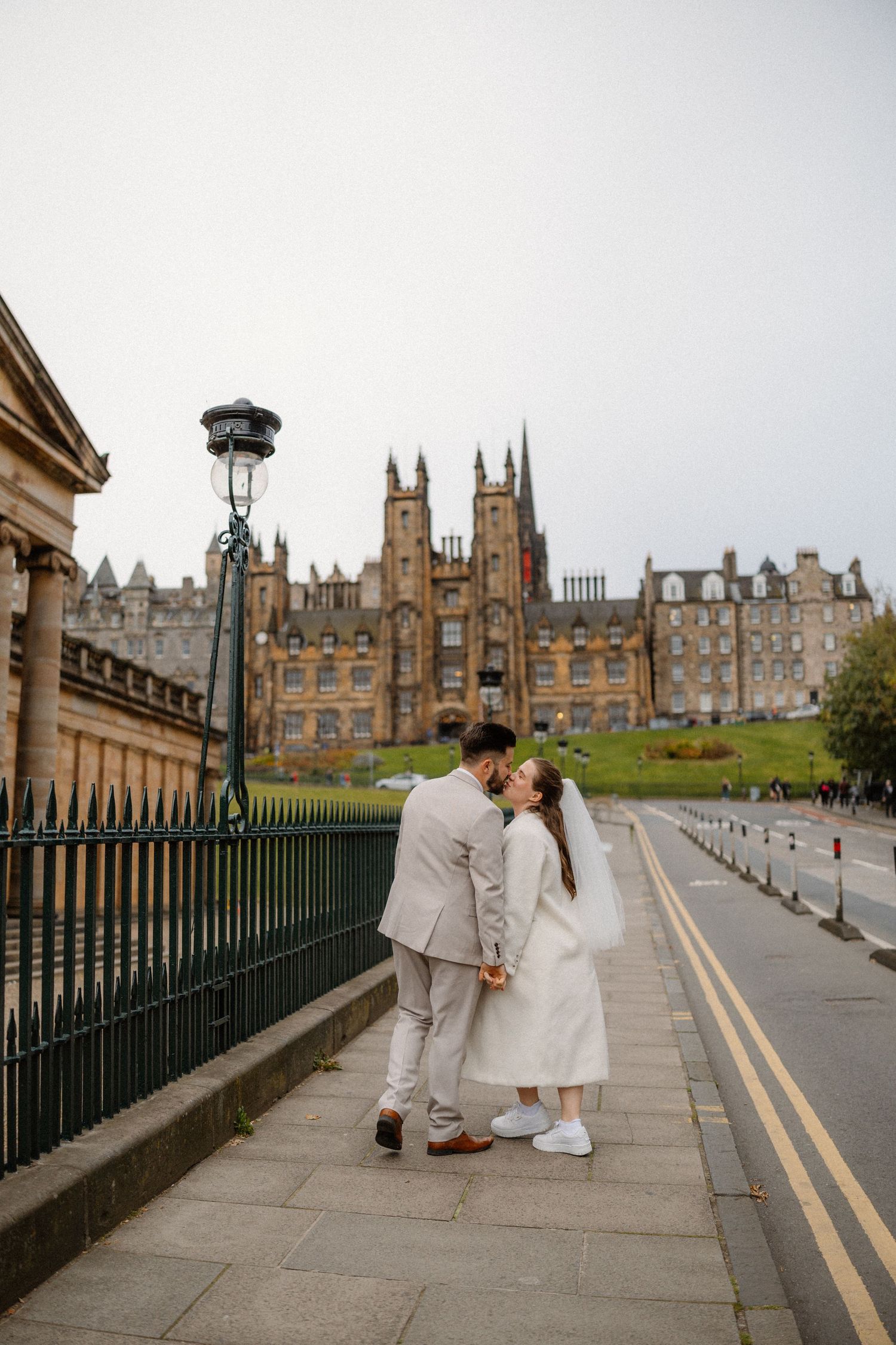 A couple in light colored outfits share a romantic moment on a sidewalk with Edinburgh's historic New College building in background.