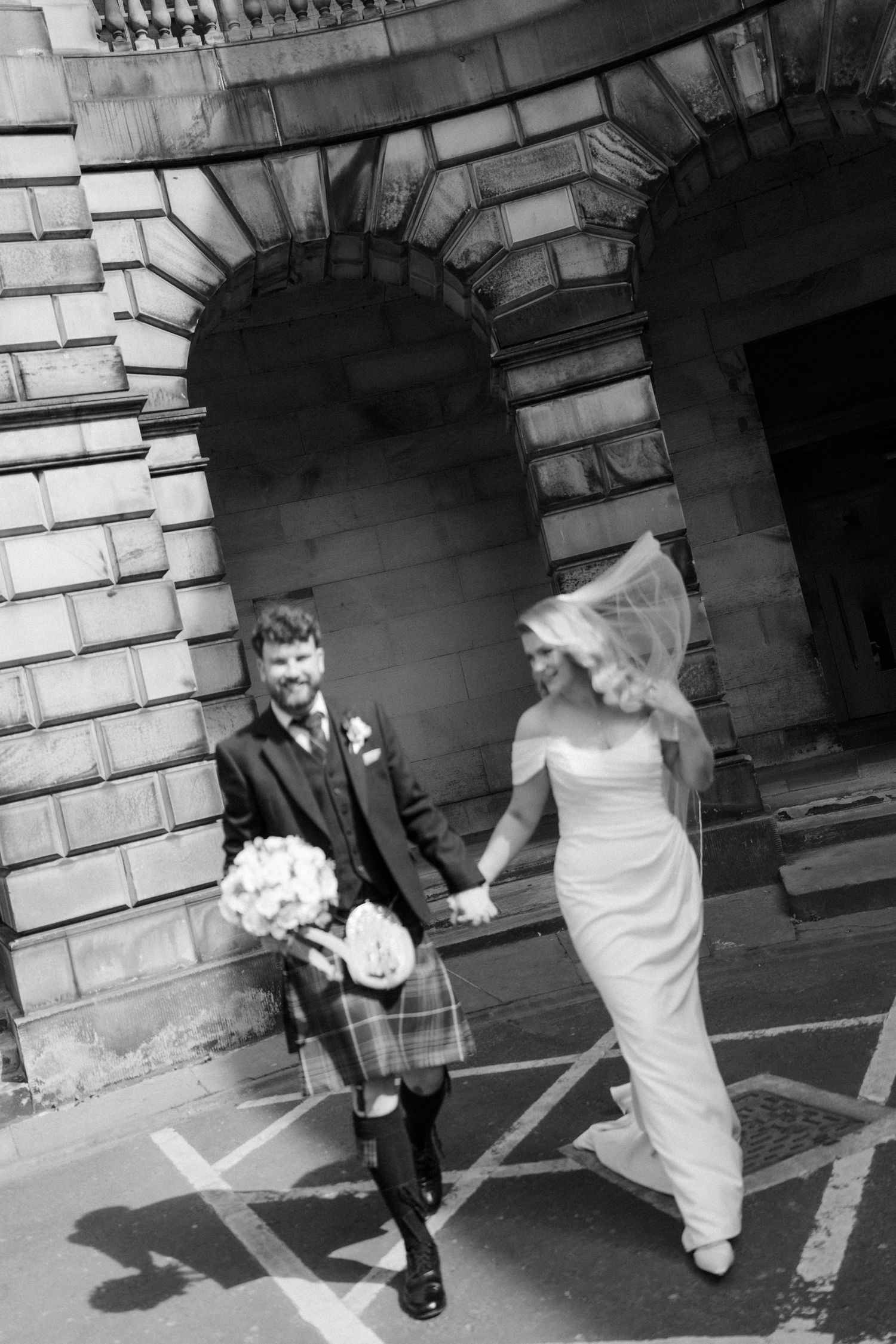 Bride and groom hold hands walking past grand stone arched building in black and white wedding photo.
