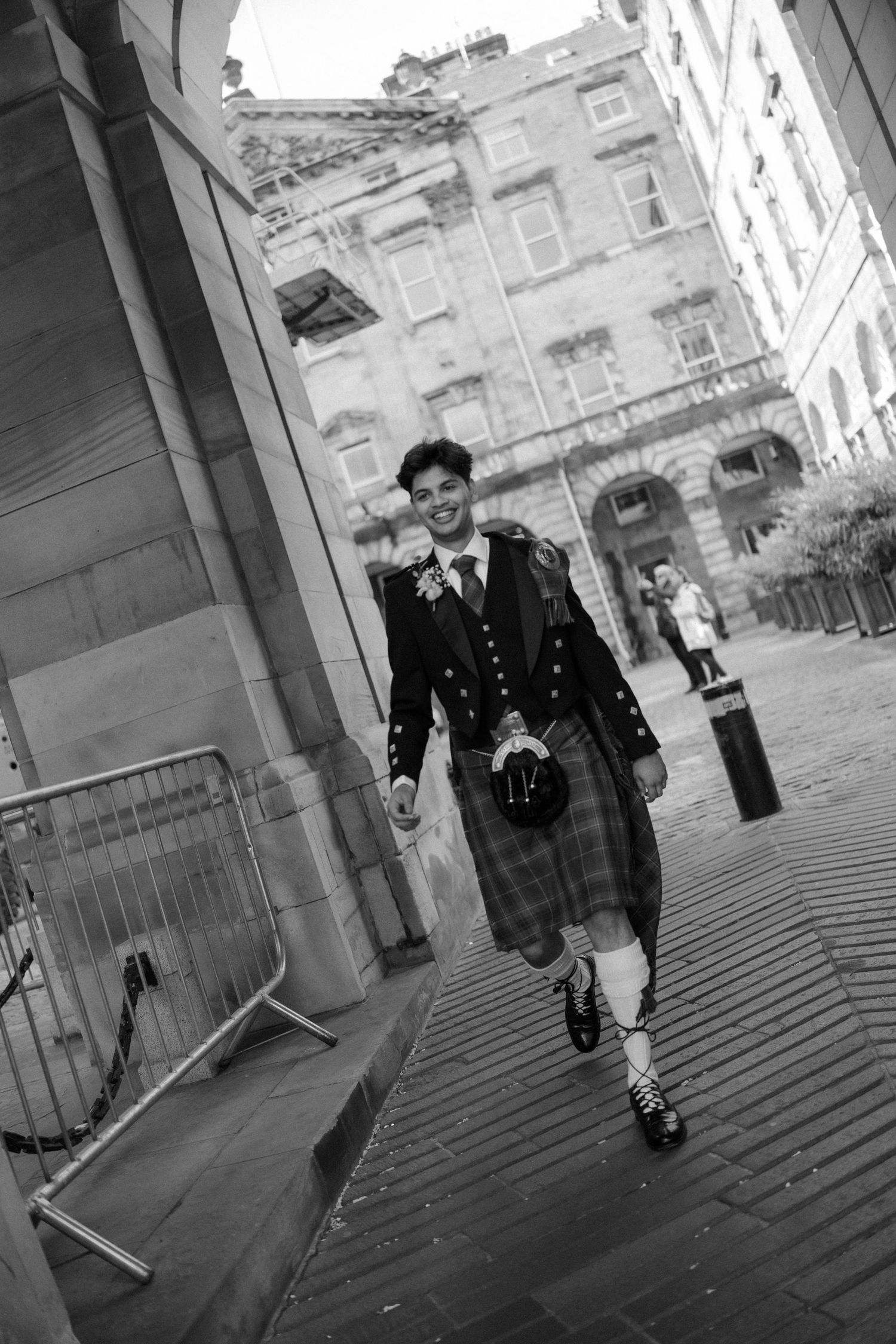 Groom in traditional Scottish kilt and jacket walking confidently through Edinburgh city streets in black and white.