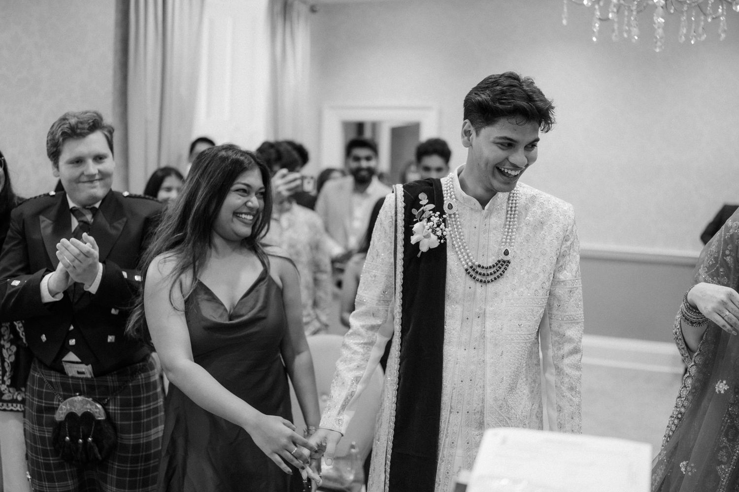 Couple holding hands smiling at Indian wedding ceremony in elegant venue, guests applauding in black and white photo.