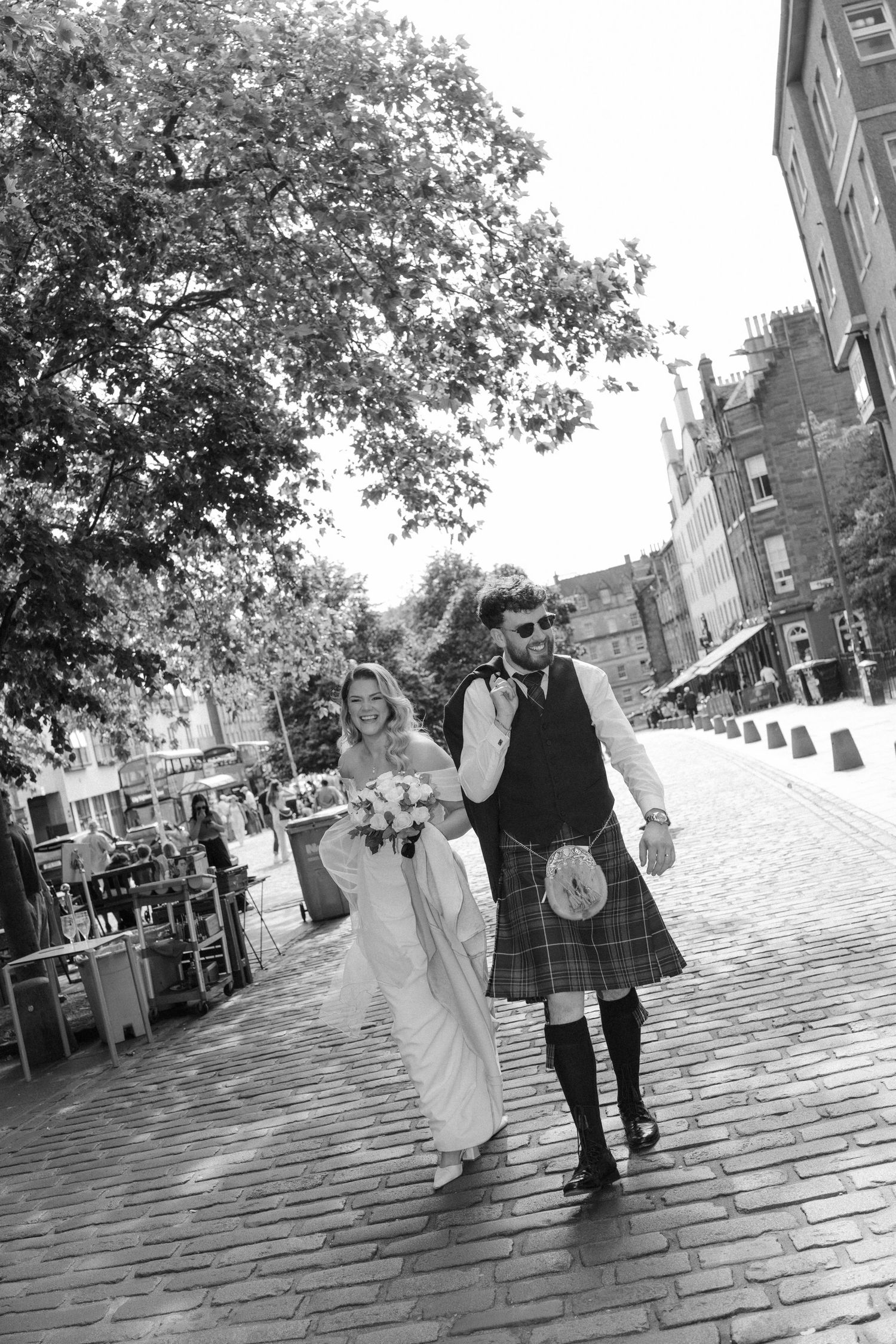 Black and white photo of a bride and groom in Scottish kilt walking on cobblestone street with bouquet.