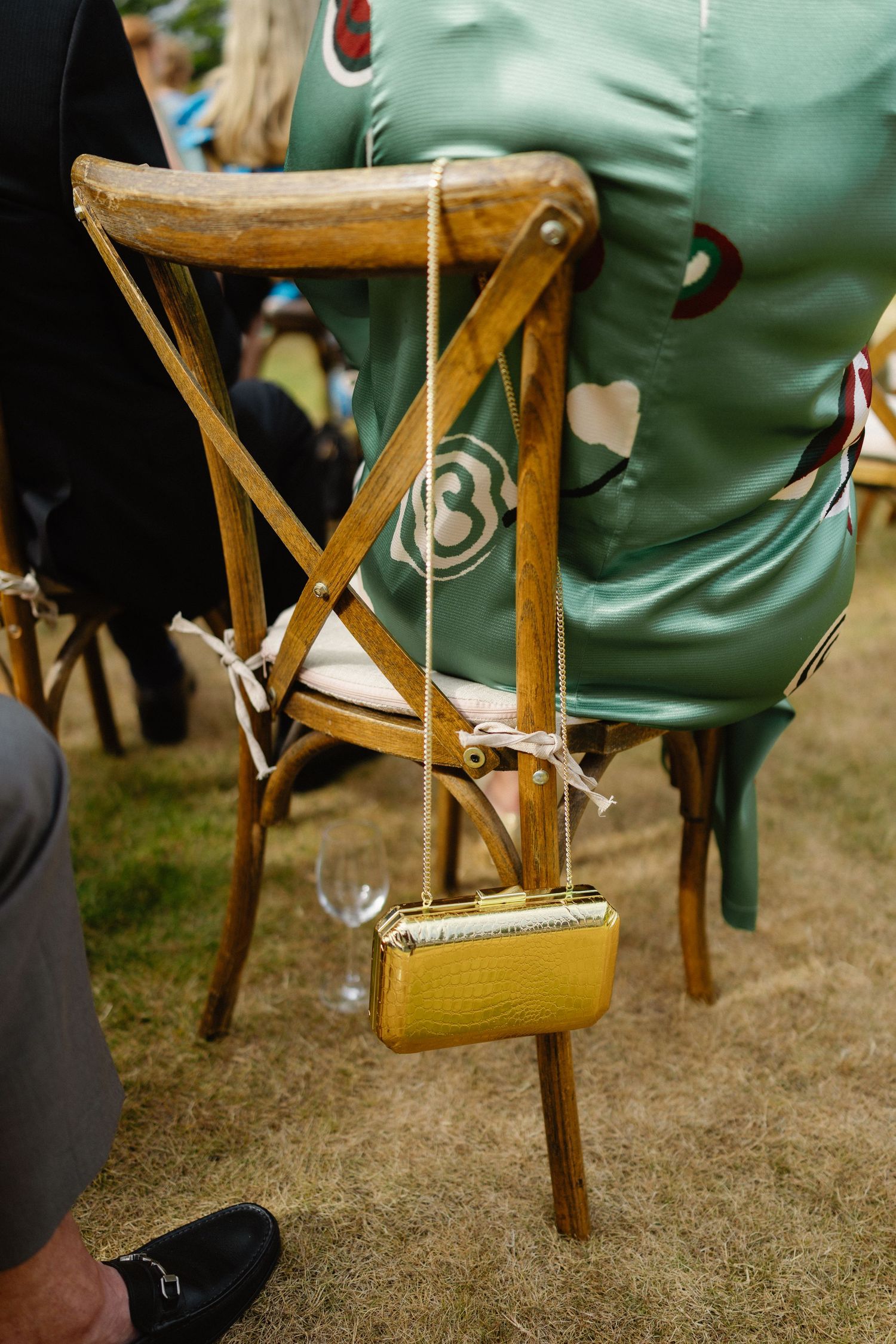 Gold clutch purse hanging on wooden cross-back chair at outdoor wedding reception.