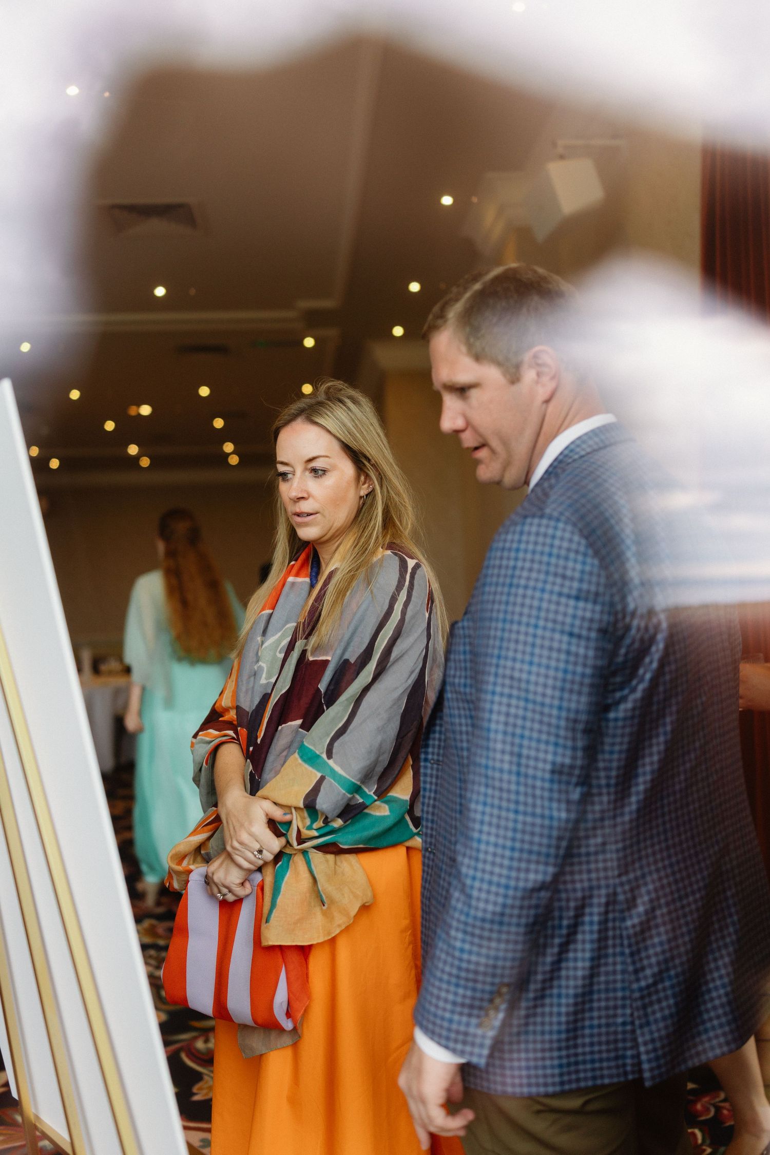 Elegant wedding guests viewing artwork at a reception, woman in colorful shawl and orange skirt with suited man.