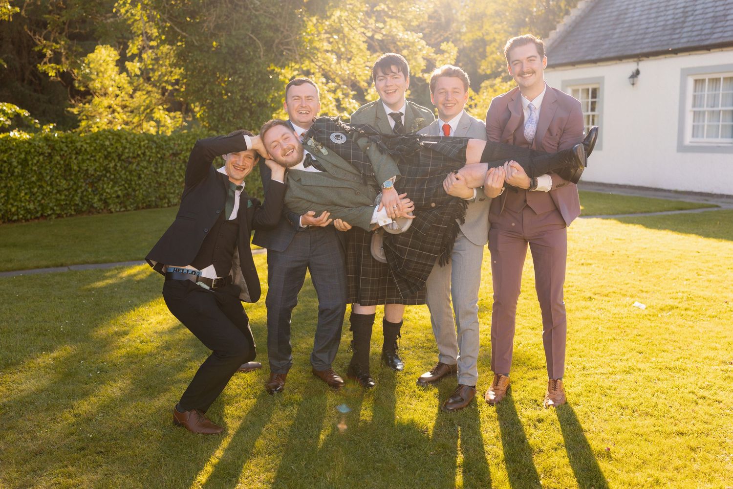 Five men in formal attire pose playfully on a sunny lawn, one wearing a kilt, at an outdoor celebration.