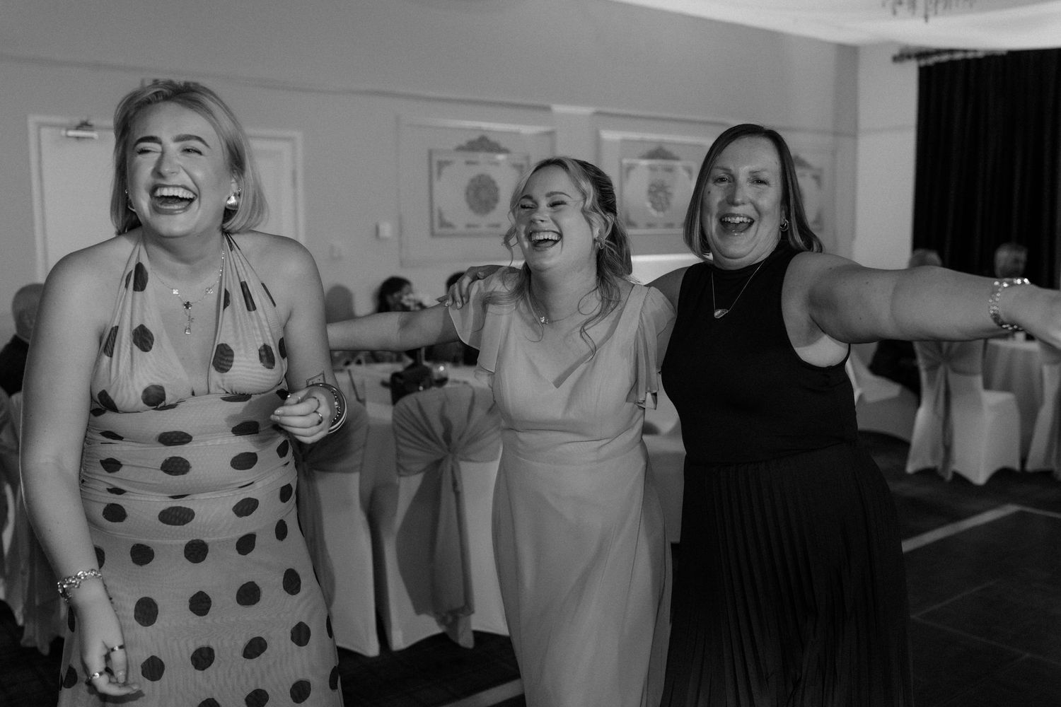 Three women laugh and dance joyfully together at a wedding reception in black and white.