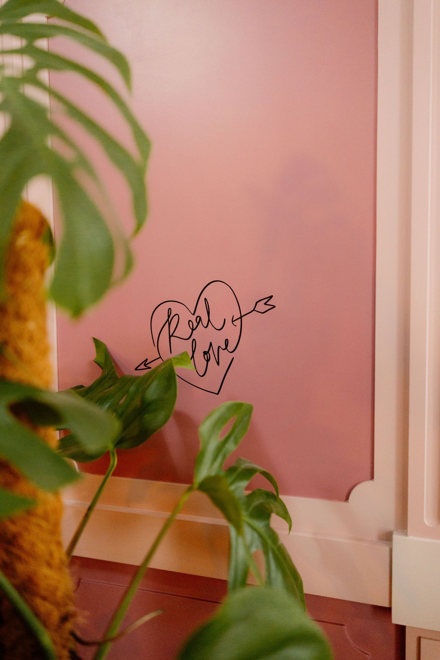 Pink wall with a heart-shaped 'Real Love' arrow sign decor, framed by lush green monstera leaves.