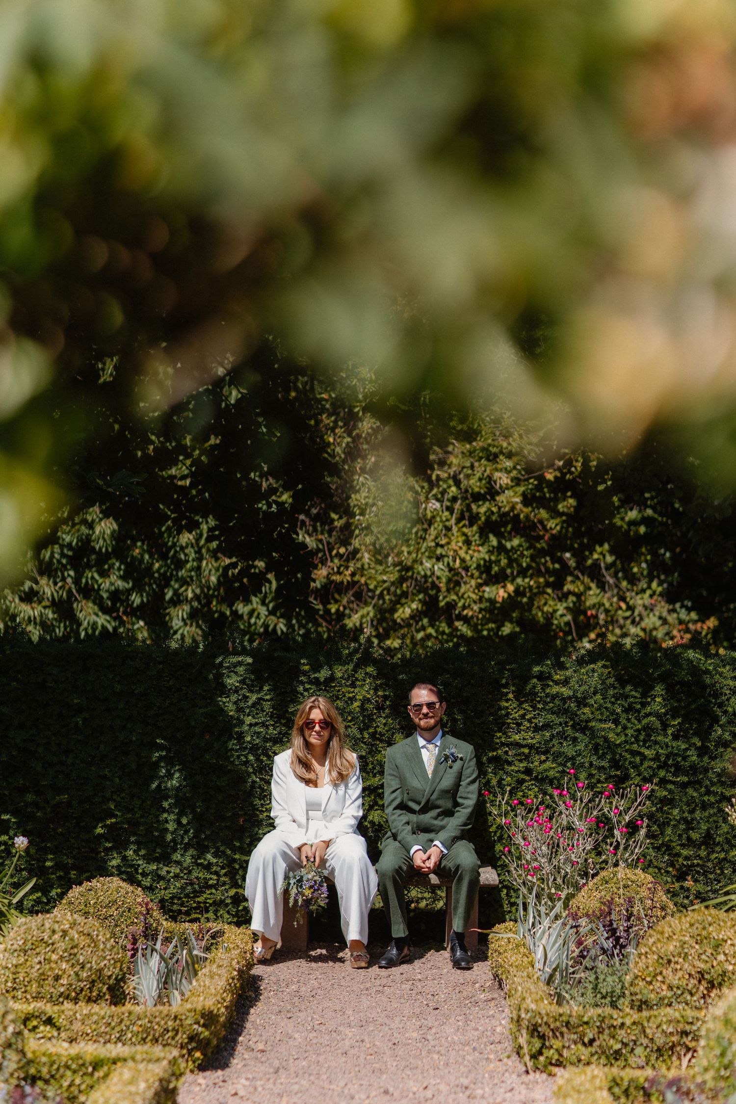 Couple sitting on garden path surrounded by lush green foliage and bushes.
