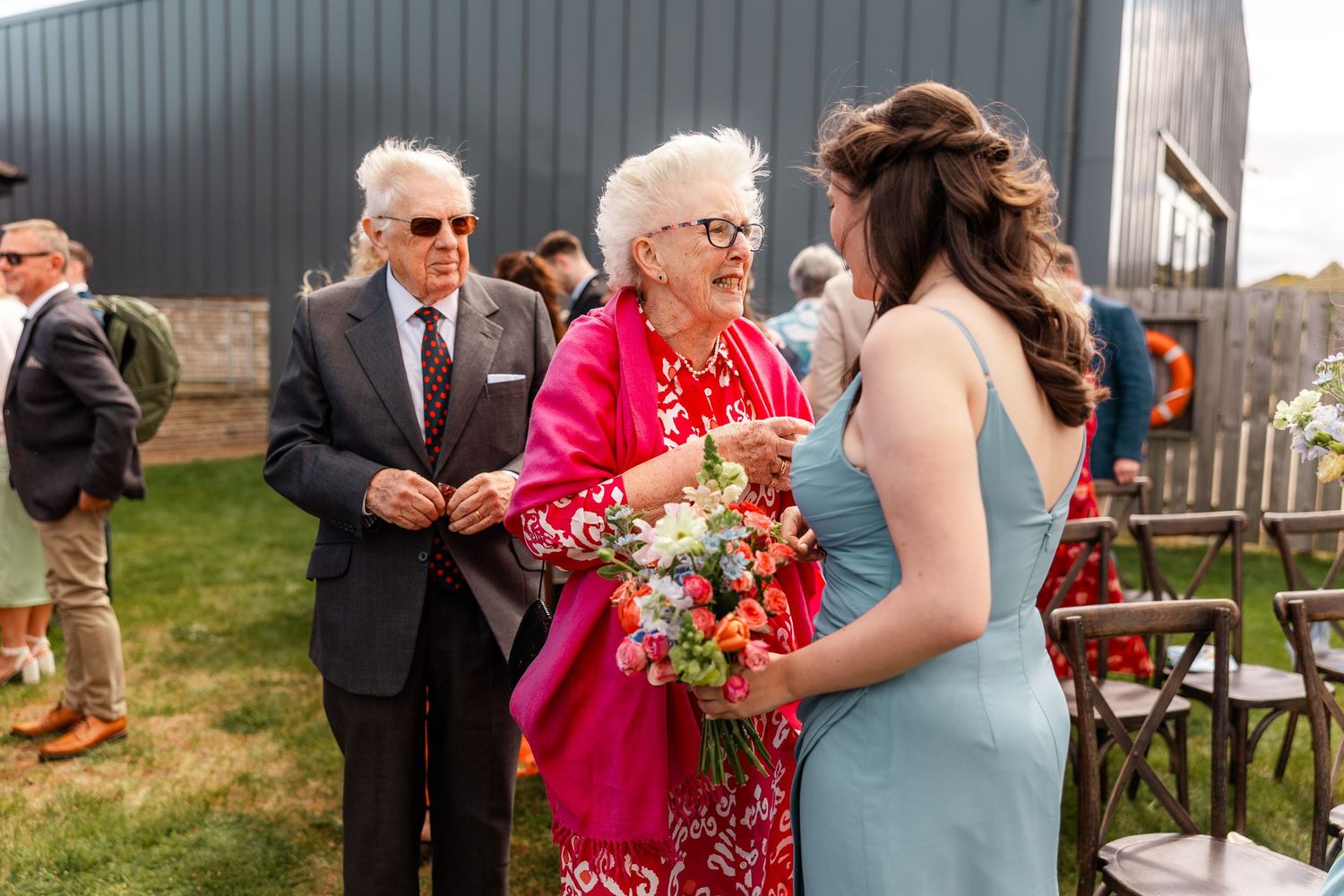 Wedding guests share a joyful moment outdoors with vibrant pink and blue attire and floral bouquets.