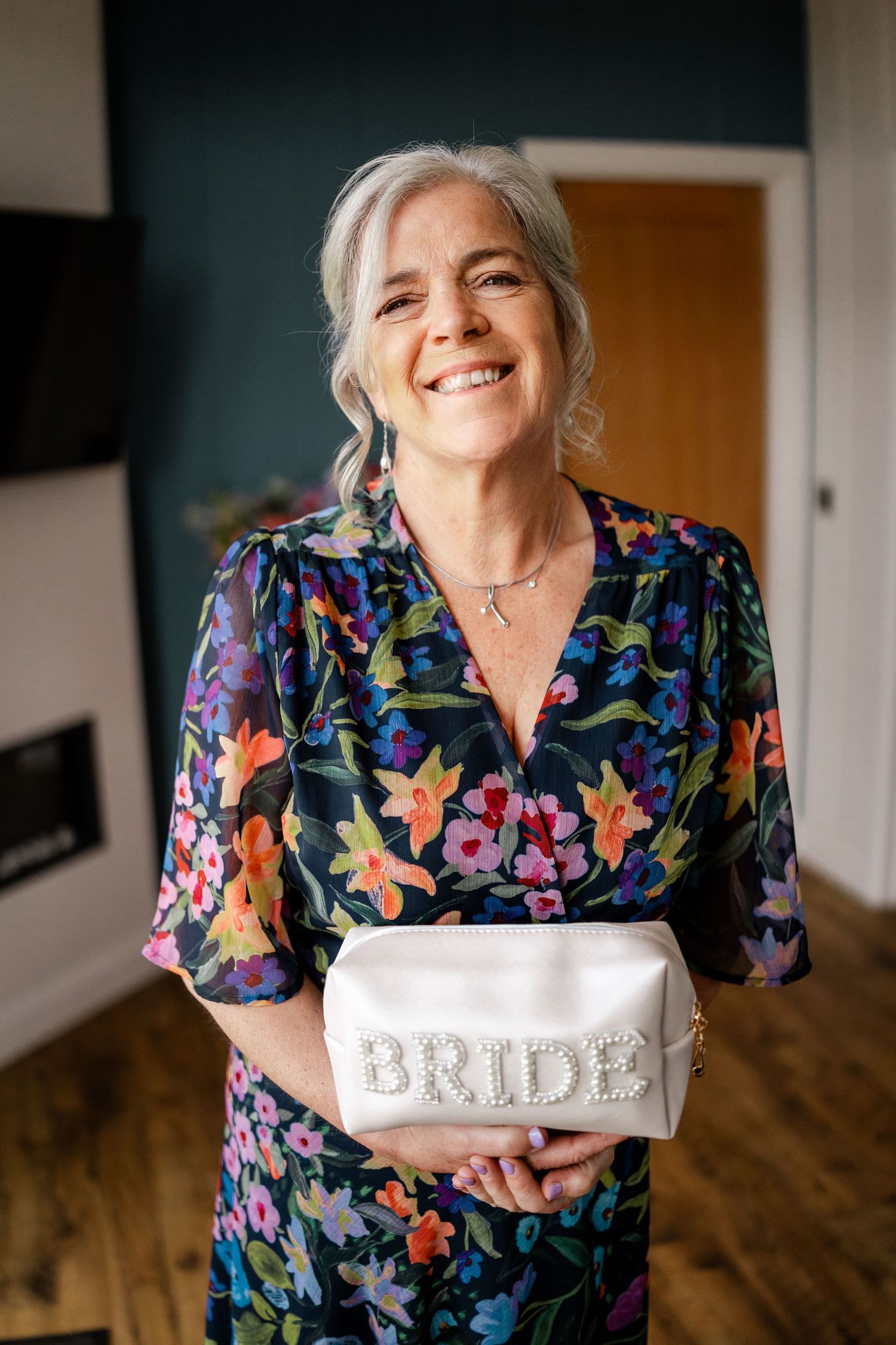 A woman in a floral print dress holds up a white cosmetic bag labeled 'BRIDE' while smiling.