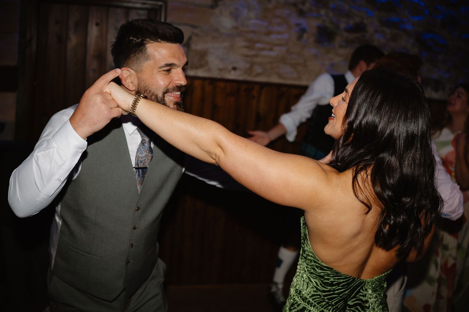 Wedding guests dancing energetically at a reception in a rustic brick-walled venue.