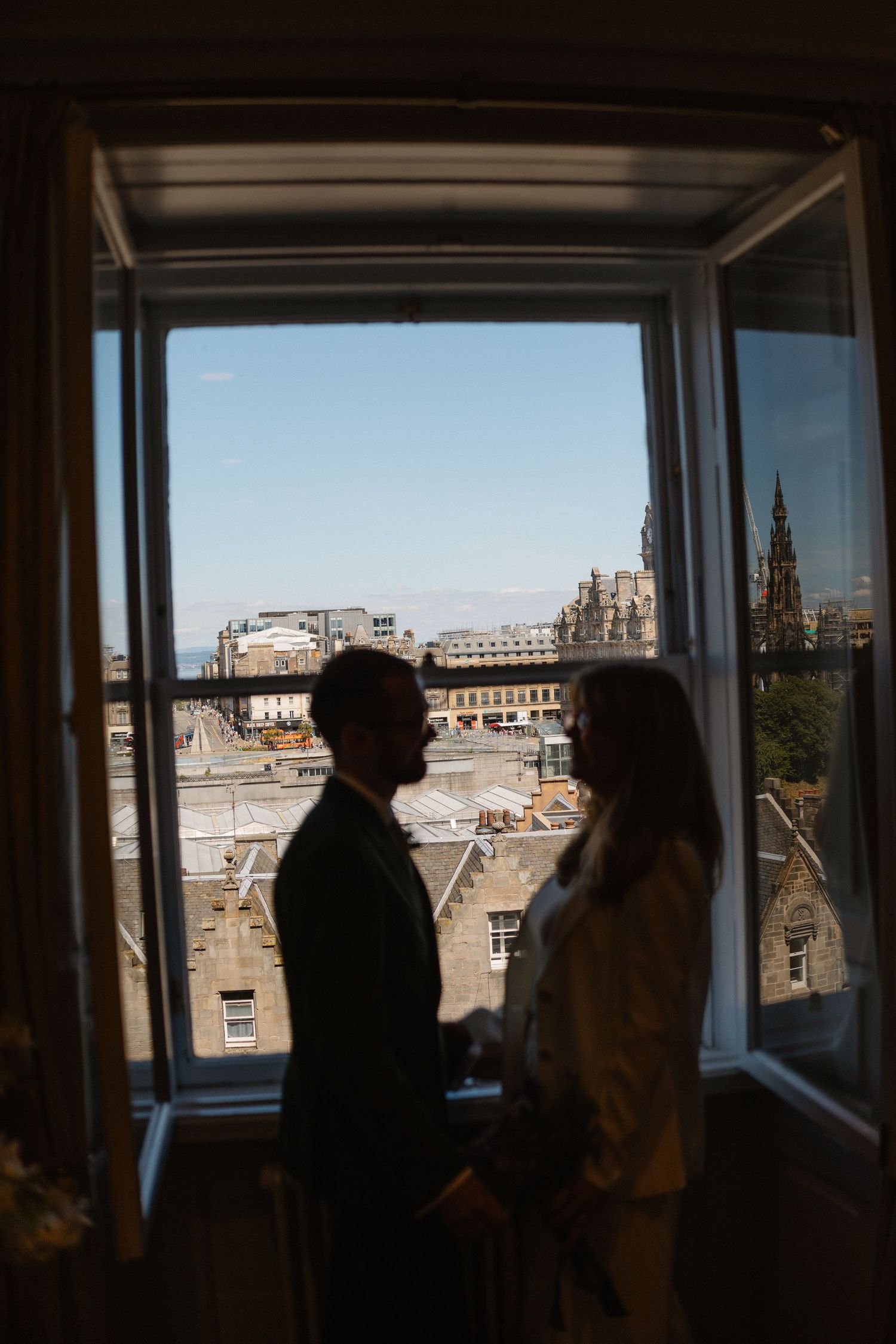 A silhouetted couple stands by a window overlooking Edinburgh's historic cityscape and Scott Monument.