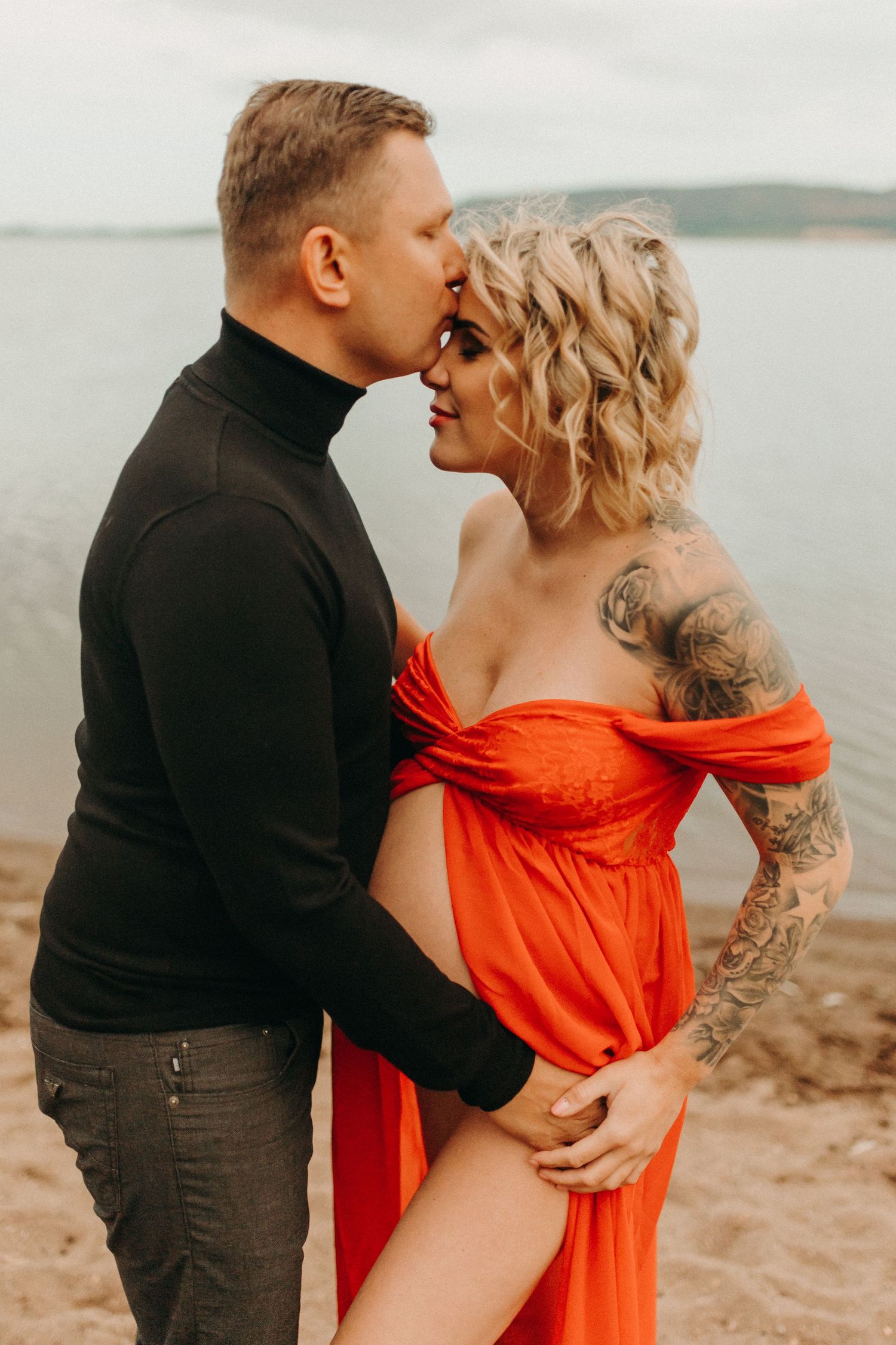 Romantic couple embraces on a beach, with woman in flowing red dress and man in black sweater sharing an intimate moment.