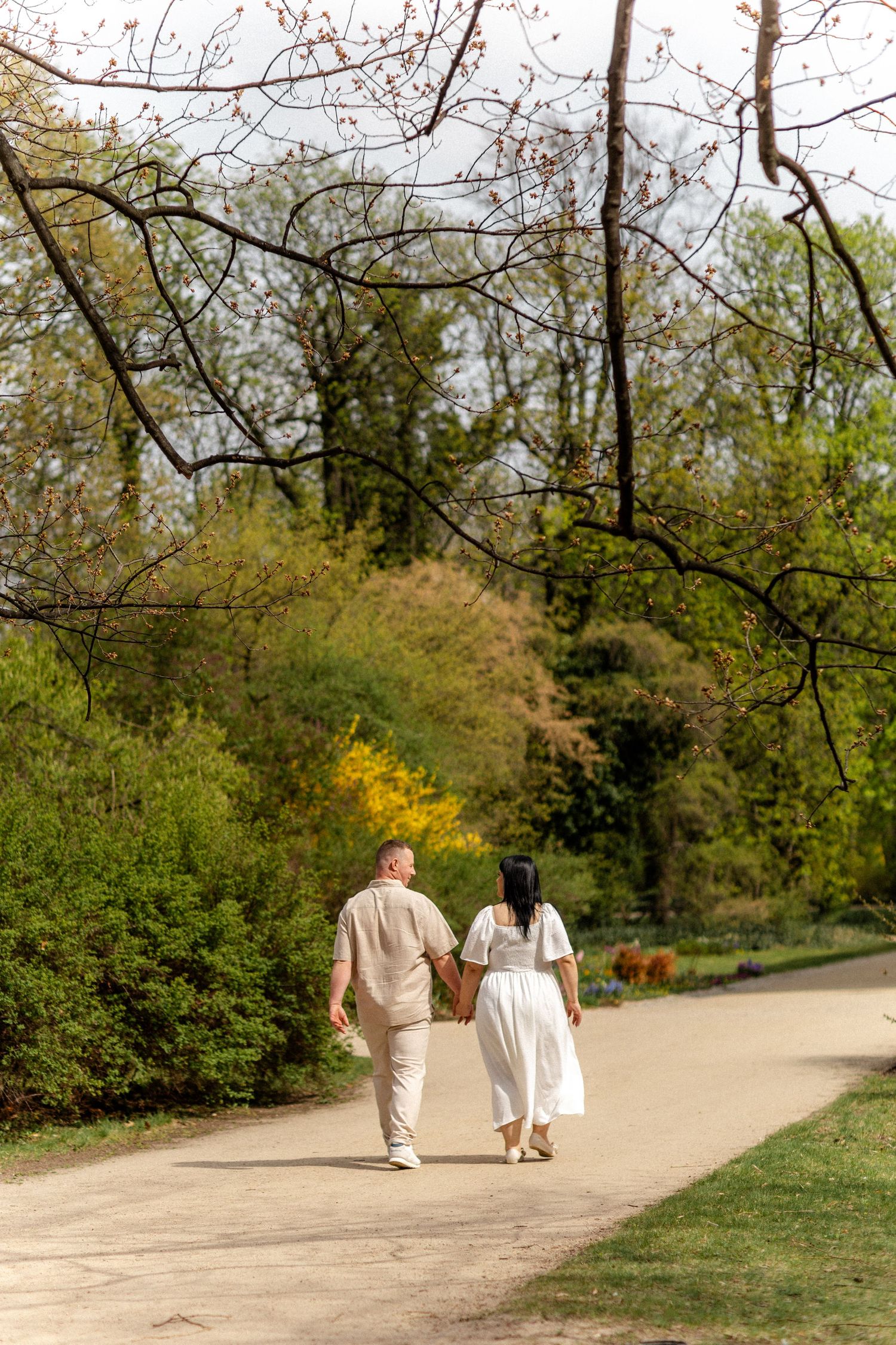 A couple walks hand in hand down a winding park path on a sunny day.
